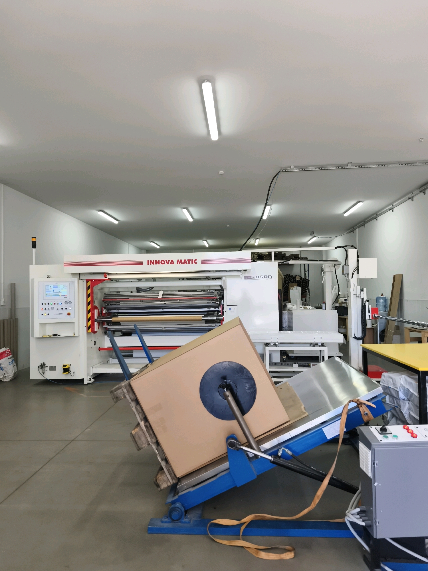 The image shows an industrial setting, likely a factory or workshop. In the foreground, a large, inclined cardboard box is secured to a blue hydraulic lift by orange straps. The box has a dark circular object attached to its surface. To the right of the lift is a grey control panel with red and green buttons. The background features a large, white industrial machine with the text "INNOVA MATIC" prominently displayed on its front. This machine appears to be a slitting or rewinding machine, with various rollers and controls visible. The floor is concrete, and the walls are white. Overhead fluorescent lights illuminate the space. The overall environment suggests an active production or processing area.