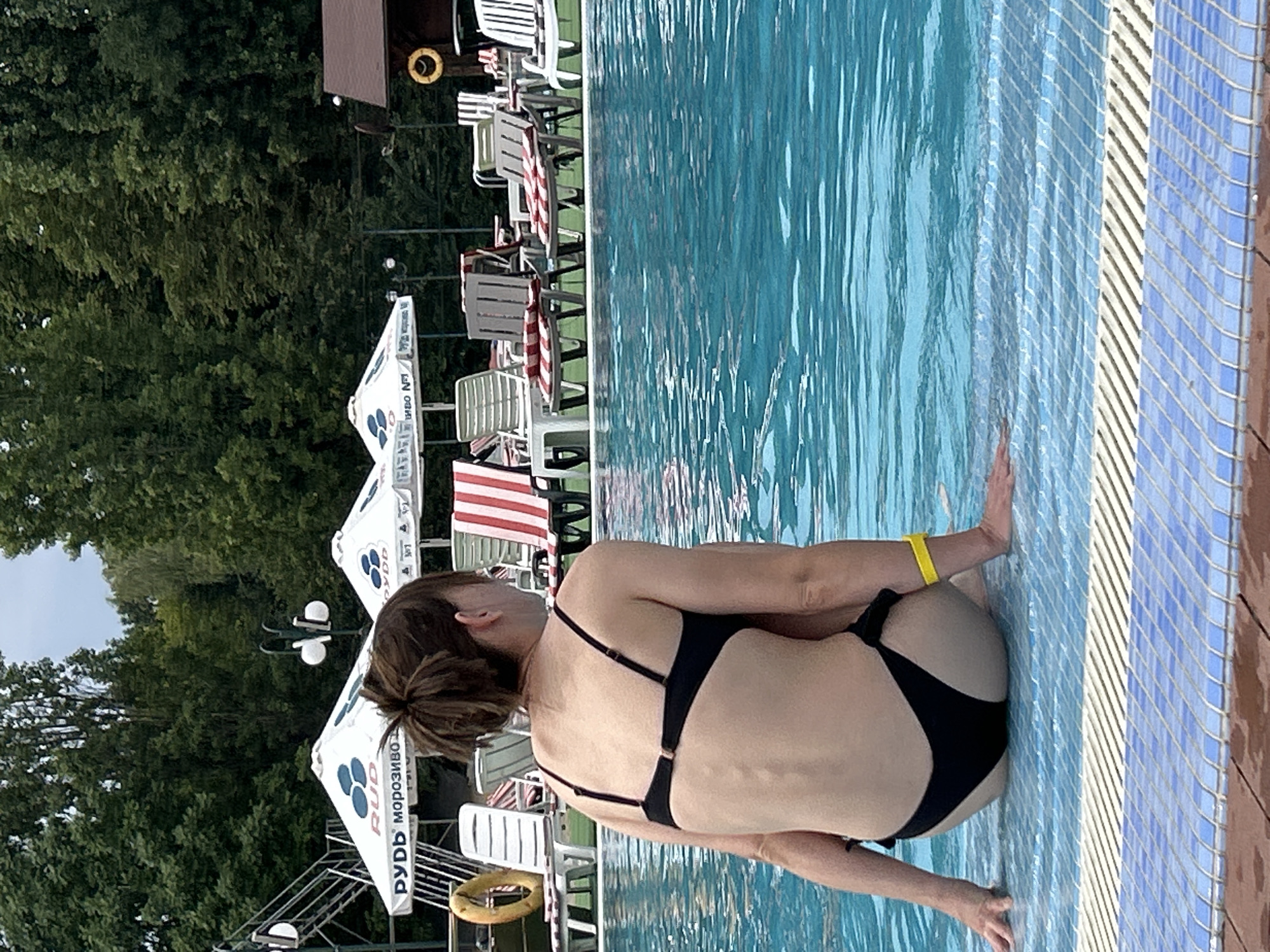 A woman in a black bikini sits on the edge of a swimming pool. She is facing away from the camera, looking towards a line of lounge chairs and umbrellas. The pool is filled with clear blue water, and the edge is lined with blue and white tiles. In the background, there are trees, a lifeguard chair, and a building. The scene appears to be set in an outdoor recreational area.