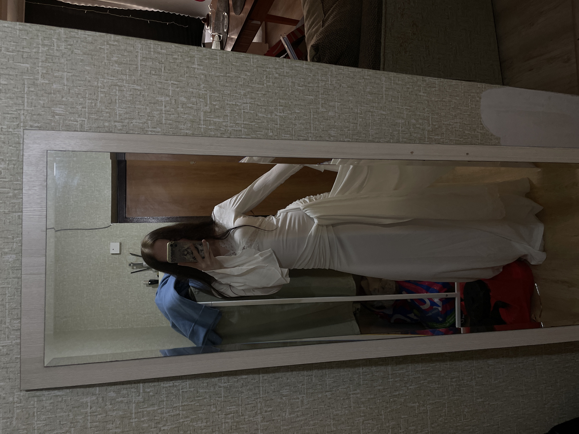 A young woman is reflected in a full-length mirror, wearing a long white dress with flowing sleeves and a sash tied at the waist. The dress has lace detailing around the neckline. She is holding a smartphone in her right hand, taking a selfie. The mirror is framed with a light wood-like material and stands against a wall with textured beige wallpaper. To the left of the woman in the reflection, there are clothes draped over a rack, and bags are visible on the floor. In the background, a wooden door is partially visible. To the right of the mirror, a glimpse of a couch and a wooden table with a teapot can be seen. The room appears to be residential, likely a bedroom or dressing area. No specific landmarks or indications of Votkinsk, Russia, are present in the image.