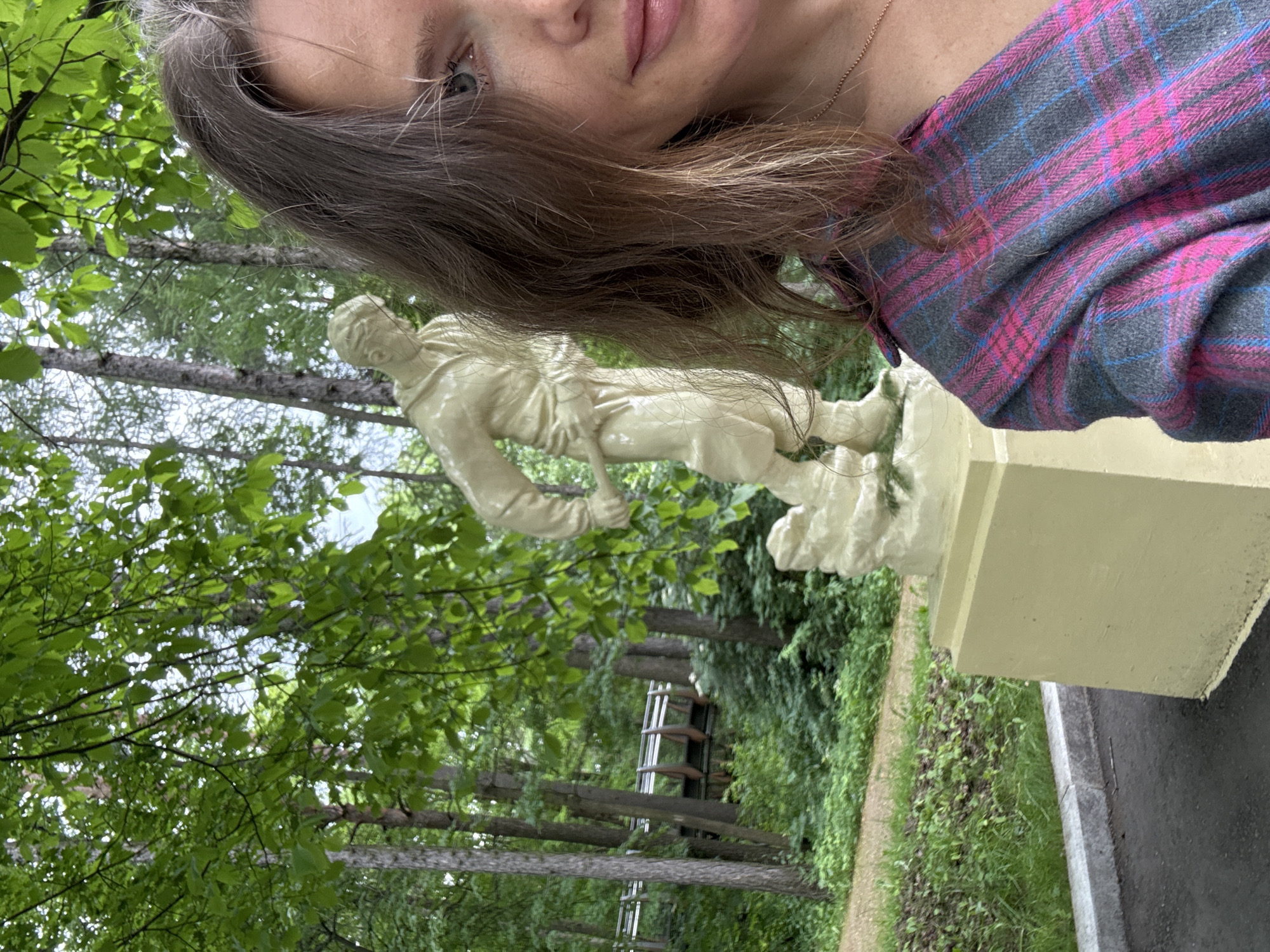 A woman is taking a selfie in front of a statue in a park. She is wearing a plaid shirt. The statue is of a man with a shovel. The park has trees and foliage. In the background, there is a bridge or walkway. The image appears to have been taken during the daytime. The location is not specified.