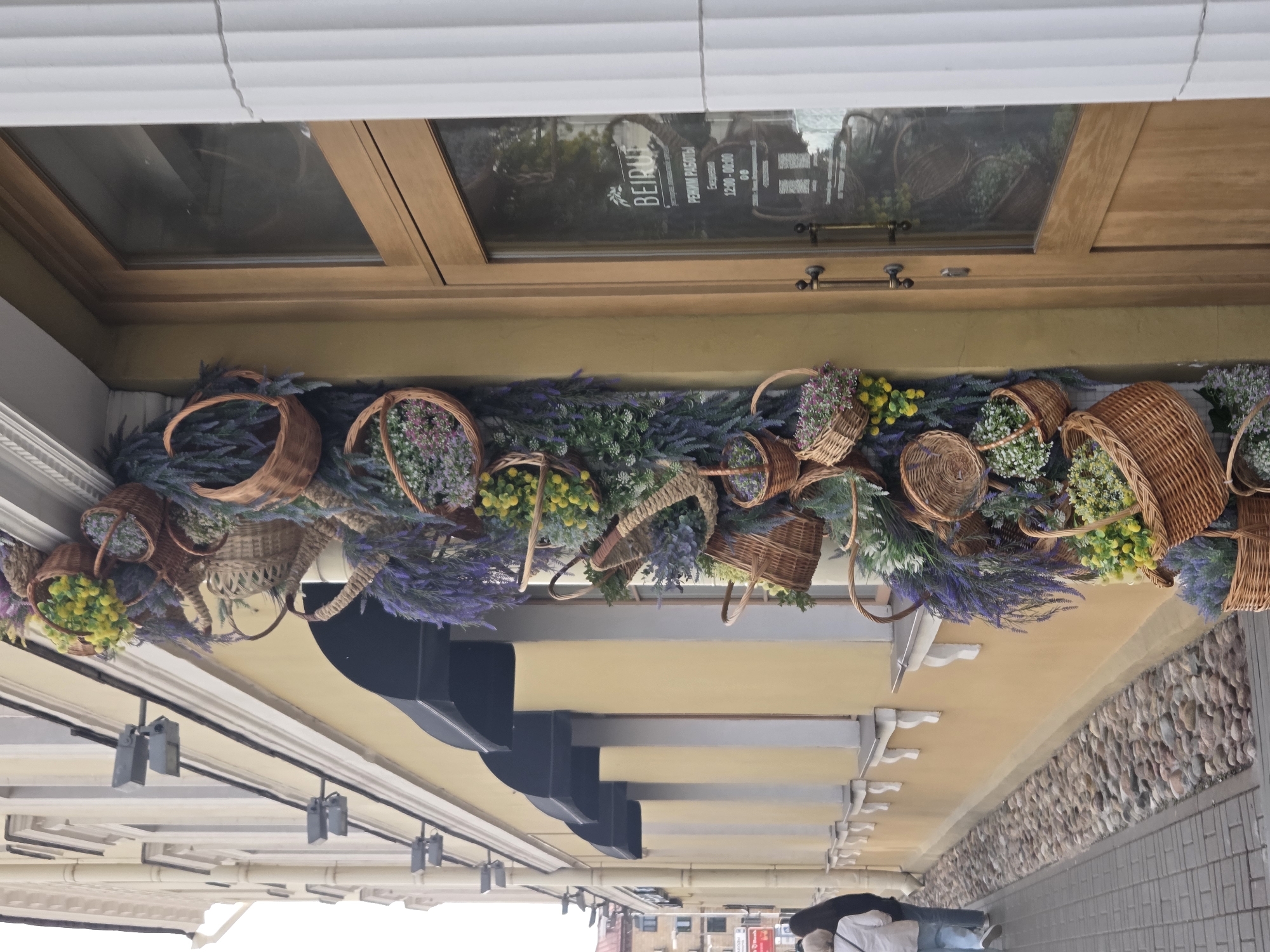 An exterior, low-angle, medium shot shows the side of a building with an elaborate floral decoration cascading down the facade, beside a wooden door with a glass panel. The building's wall is painted a pale yellow, and several dark awnings are visible above windows. The floral decoration consists of numerous woven baskets of various sizes, filled with artificial flowers and foliage in shades of purple, green, and yellow. The baskets are arranged in a spiraling pattern along the wall. The wooden door is to the right of the floral display, and its glass panel reflects the surroundings. Text is visible on the glass, indicating "BEIRUT" and "РЕЖИМ РАБОТЫ" (working hours). A white fluted column is partially visible to the right of the door. In the foreground, to the left, a grey paved sidewalk is bordered by a narrow strip of river stones. Two individuals, one in a white jacket and blue jeans, and another in a black jacket, are walking away from the camera on the sidewalk. The overall scene suggests an outdoor storefront or establishment in an urban setting.