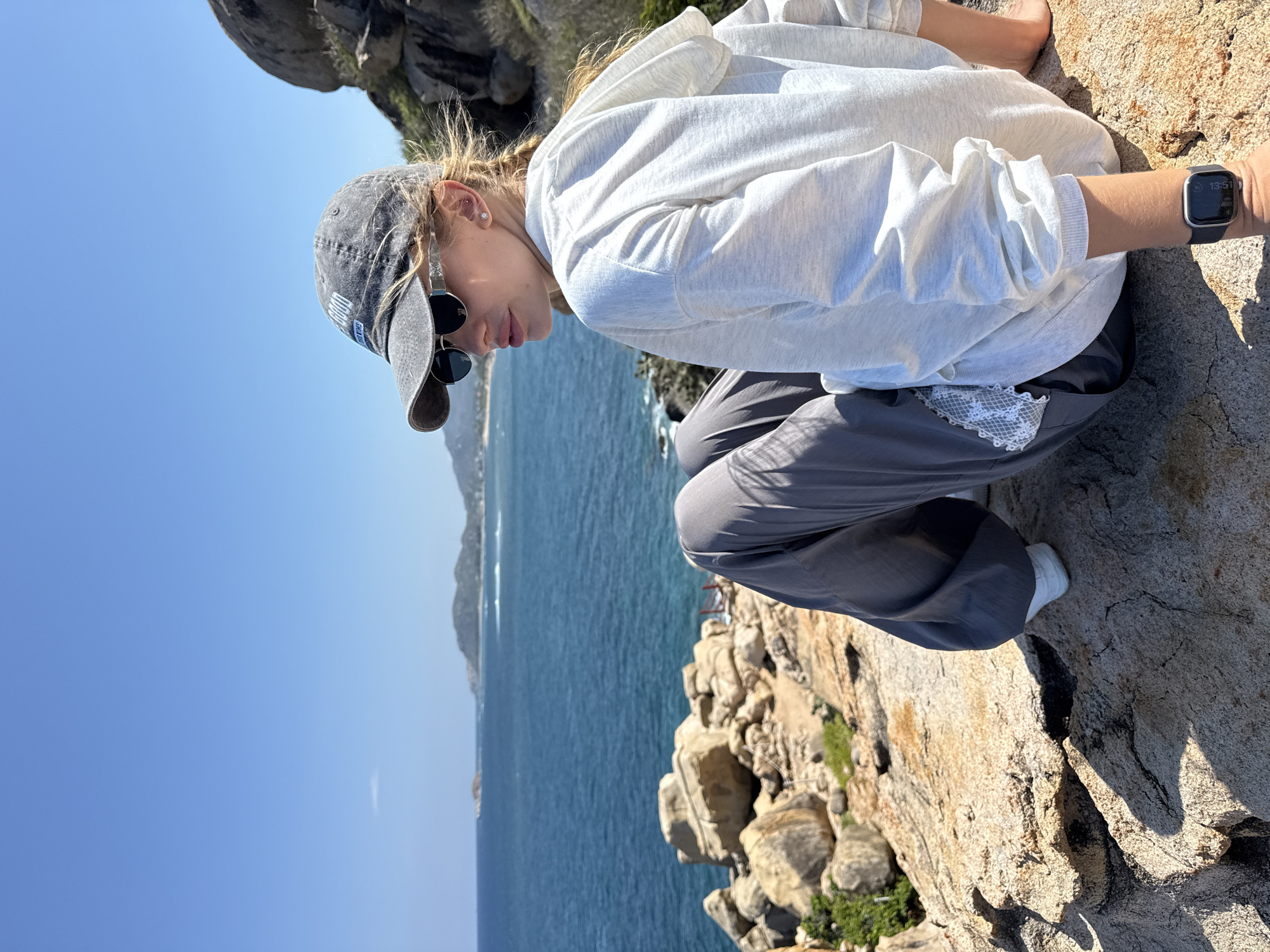 A young woman is crouching on a rocky outcrop overlooking the ocean. She is wearing a light grey hooded sweatshirt, dark grey pants, a grey baseball cap, and dark sunglasses. She is also wearing a watch on her left wrist. The ocean stretches out to the horizon, with a mountain range visible in the distance. The sky is clear and blue. The location is Vinh Hai, Vietnam, judging by the landscape.