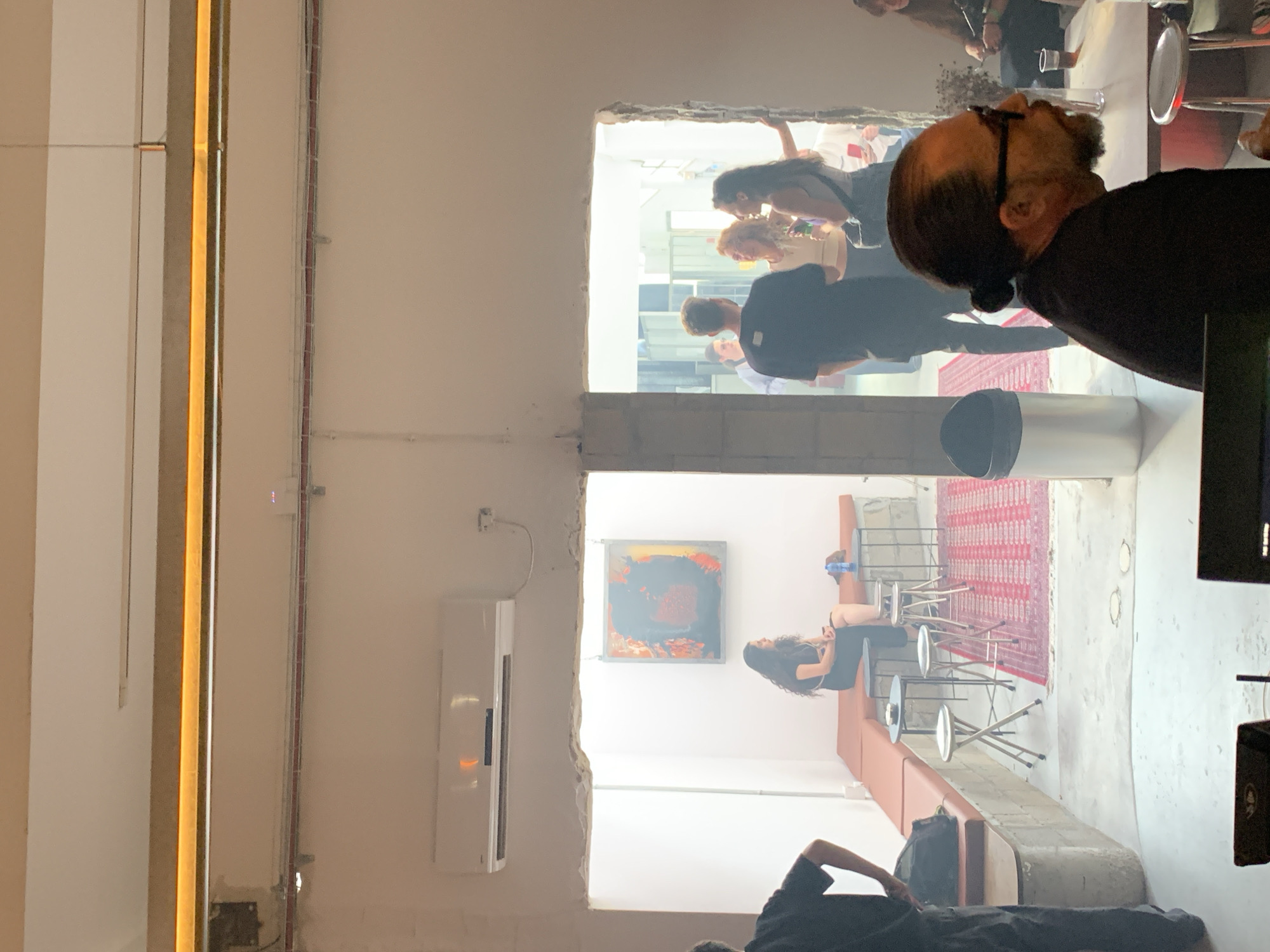 The image presents an interior view within a structure, likely located in Tel-Aviv, Israel. Two rectangular openings divide the scene into distinct sections.
The upper opening reveals a gathering of people, presumably socializing. Several individuals stand or lean against surfaces, suggesting a casual atmosphere.
The lower opening shows a seating area with a red rug. A woman is seen sitting on a stool. In the foreground, another person is sitting down.
Architectural details within the building include a long light fixture, electrical conduits running along a wall, and an air conditioning unit. Additionally, artwork hangs on the wall.