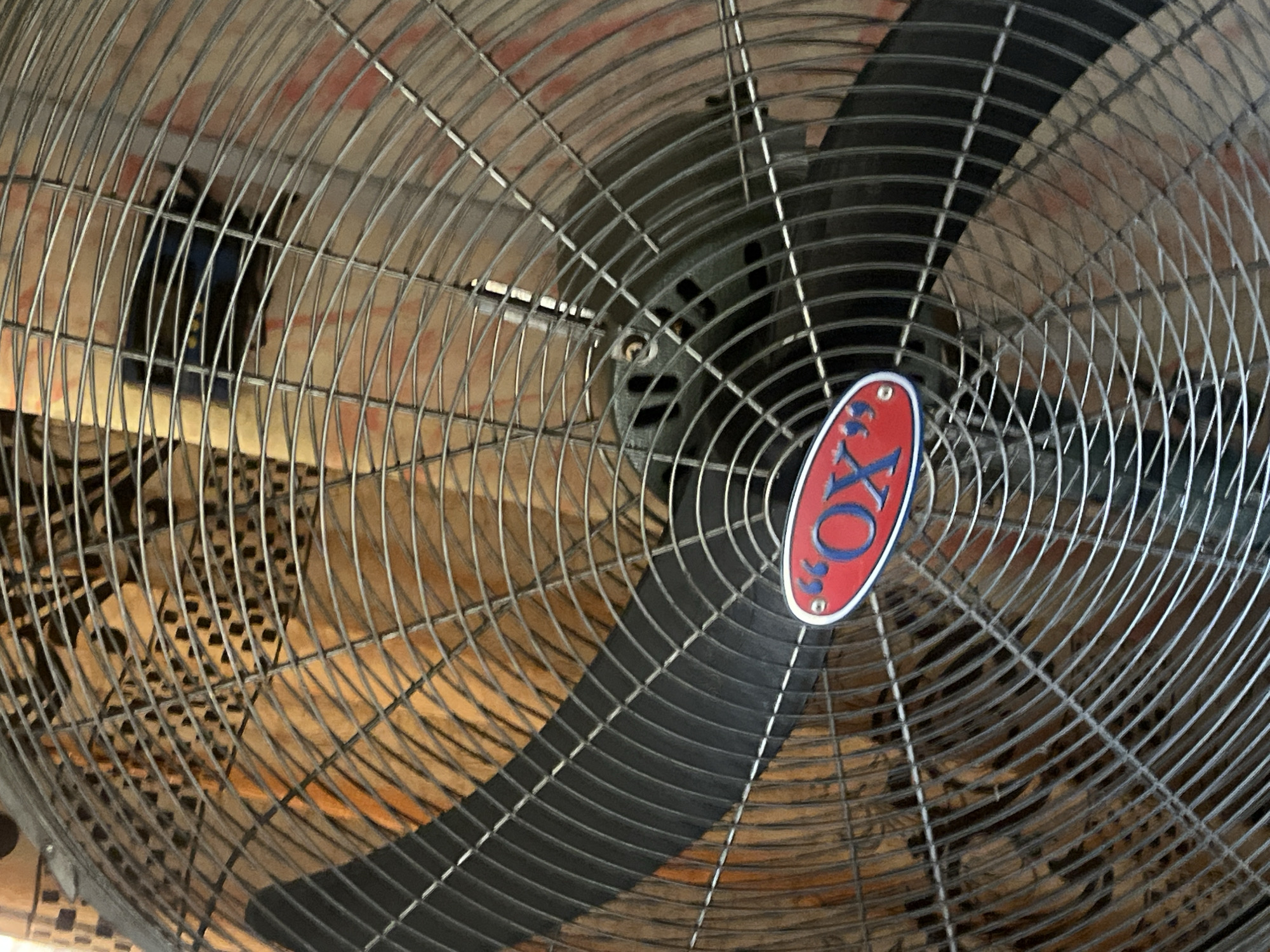 The image shows a close-up of a metal oscillating fan with a black blade and a red and blue oval logo that reads "OX". The fan grille is made of thin metal wires. In the background, there is a blurred image of what appears to be wooden planks and a patterned fabric. The fan is positioned indoors, with a wall visible behind it. There are no people or animals present in the image. The overall impression is a still life of a functional object in an indoor setting. Without further information, it is not possible to determine the specific location in Jalingo, Nigeria.