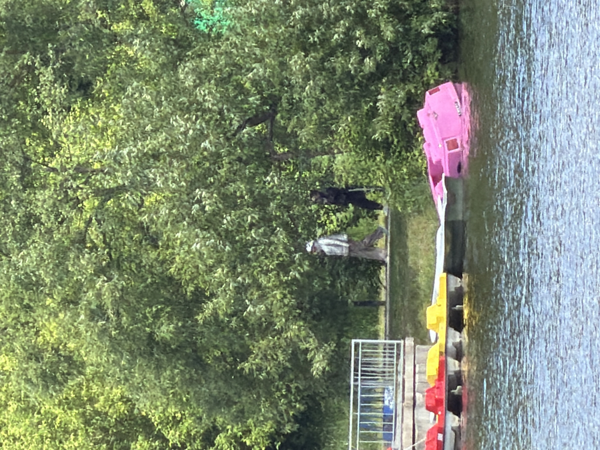 The scene depicts a lake in Malakhovka, Russia, bordered by lush green trees. A dock extends into the water, where a collection of colorful paddle boats, predominantly pink, yellow, and red, are moored. Beside the dock, a metal railing stands. In the background, two individuals are visible on the grassy bank. One is dressed in dark clothing, while the other is wearing lighter attire. The overall impression is one of a peaceful, recreational area.