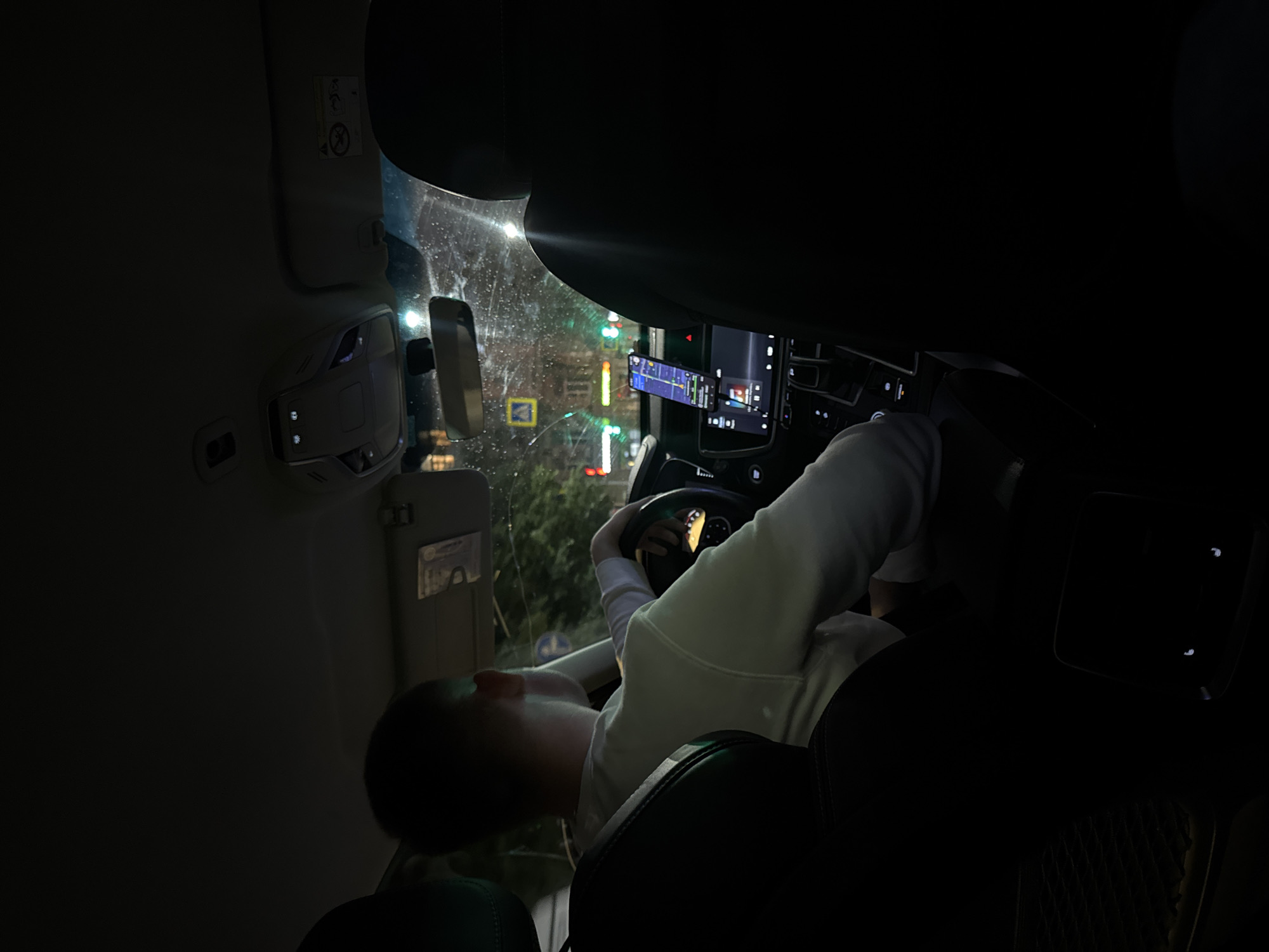 The image shows the interior of a car at night, seen from the passenger seat. A person is driving, with their back to the camera. They are wearing a white sweatshirt and have their hands on the steering wheel. The dashboard has a central screen displaying navigation and music information, and a phone mounted on a holder. The windshield shows streetlights and buildings outside. The scene appears to be in a city, possibly in traffic, as suggested by the lights and the navigation display.