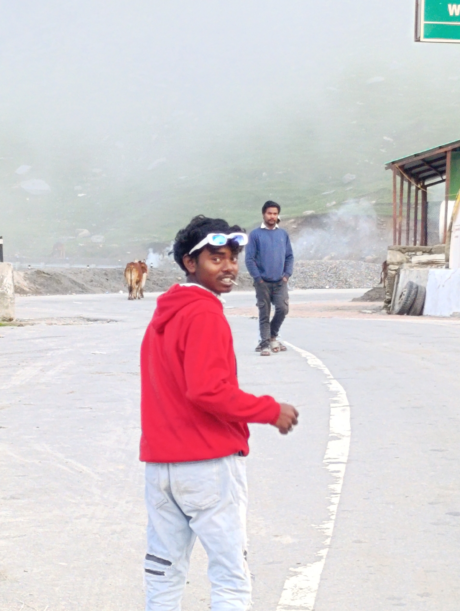 In Marhi, India, two young men are captured in a photographic moment on a paved road. The foreground is dominated by a man wearing a red hooded sweatshirt and light-colored, distressed jeans. He is turned towards the viewer, offering a slight smile, with a cigarette held between his lips and white-rimmed sunglasses perched on his head. His posture suggests a relaxed, informal demeanor.
Further back on the road, another man stands with his hands in the pockets of his dark trousers. He is wearing a blue crew-neck sweater over a white collared shirt, and his gaze is directed away from the camera.
To the left of the men, a light brown cow stands facing away from the camera, its presence indicating a rural or semi-rural setting. In the background, a gentle slope rises, partially obscured by a light, hazy atmosphere. A small, rustic structure with a sloped roof is visible on the right side of the image, with a green sign overhead. Two tires are stacked near the structure. The overall environment suggests a mountainous region, possibly a roadside stop or a village in the Himalayas.