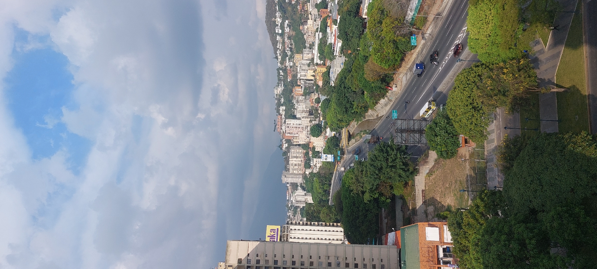 This high-angle, panoramic view captures a bustling urban landscape, likely in Caracas, Venezuela, given the context. The scene is dominated by a clear, partly cloudy sky, with patches of bright blue visible through soft, white and grey clouds.
In the mid-ground, a multi-lane highway stretches across the image, populated by various vehicles including cars, trucks, and motorcycles. The highway is flanked by lush green trees and interspersed with small, green road signs, though their text is illegible. Alongside the road, there are paved walkways and manicured green spaces, suggesting public park areas.
Beyond the highway, the city unfolds with numerous buildings of varying sizes and styles, climbing up the sides of green, forested mountains in the distant background. These structures appear to be a mix of residential and commercial developments, forming a dense urban tapestry against the natural elevation. The mountain peaks are hazy, blending into the distant horizon under the cloudy sky.
In the foreground, closer to the viewer, more urban infrastructure is visible, including apartment buildings and other commercial structures. A notable detail is a large, yellow sign atop a multi-story building that clearly reads "aka." There are also more trees and a visible patterned walkway in this section.
The image depicts a typical daytime scene, with bright, diffused light indicating either morning or afternoon. The overall atmosphere is one of active urban life set against a dramatic mountainous backdrop. No specific individuals or strong emotions are discernible, presenting a factual snapshot of the city environment.