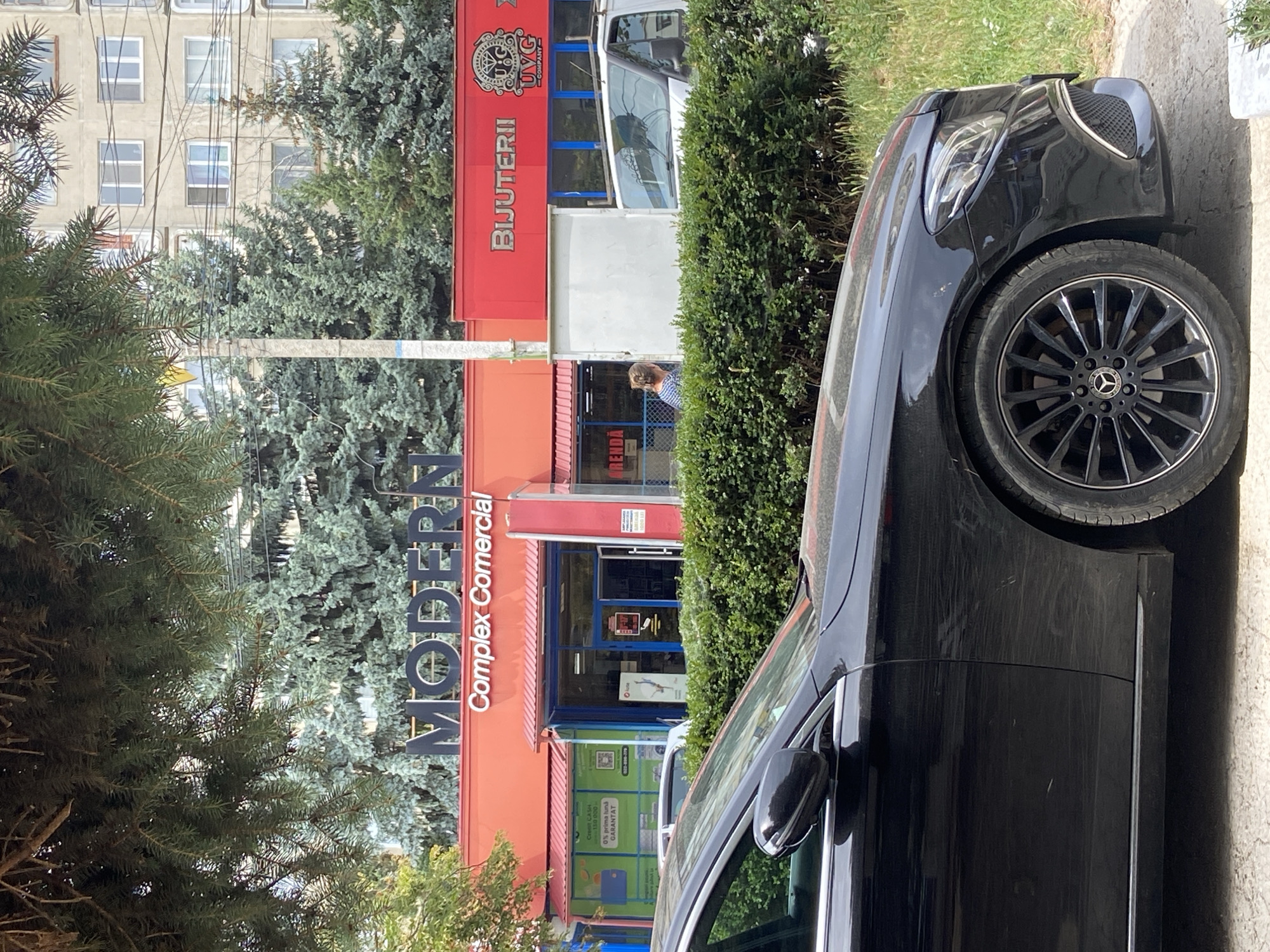 A black Mercedes-Benz sedan is parked in front of a commercial building. The building has an orange facade with the word "MODERN" in large, dark lettering at the top. Below "MODERN" is smaller text that reads "Complex Comercial". To the right of "MODERN", the word "BIJUTERII" is visible in red lettering, with a circular logo above it. There is a white van parked to the right of the building. A person is visible through the building's glass doors, appearing to be looking out. A hedge runs along the front of the building, partially obscuring the lower portion of the van and the Mercedes. Lush green trees, including some with blue-green needles, are behind the commercial building. The sky is not visible in the frame. The ground in front of the car appears to be paved.
