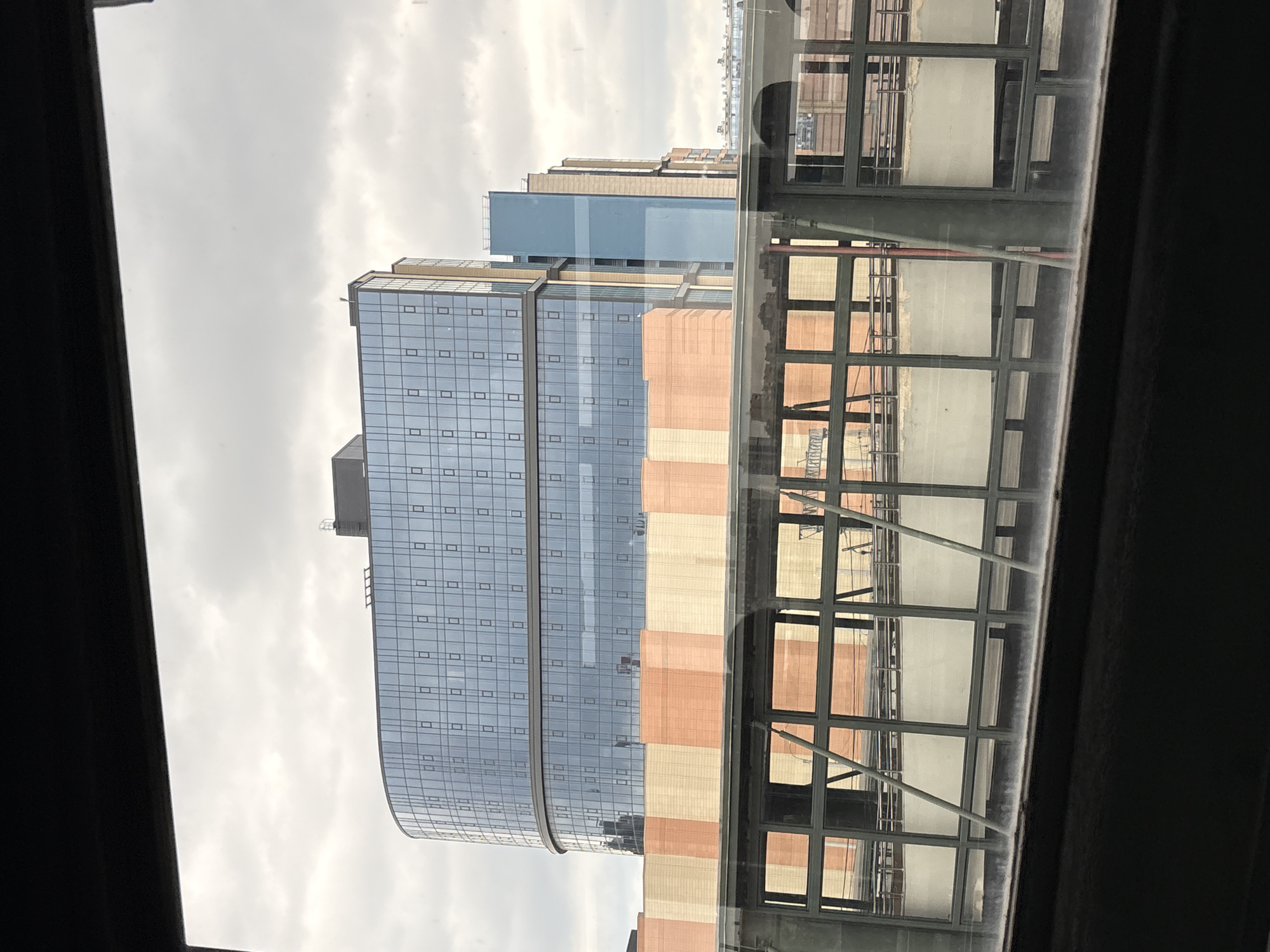The image shows a view of modern buildings, possibly a complex of offices or residential apartments, seen through a window. The buildings feature extensive glass facades with a grid pattern of small, rectangular windows. The exterior of one building is accented with alternating bands of light beige and salmon-colored panels. A covered walkway or skybridge with a steel frame and glass panels runs in front of part of the buildings, reflecting the architectural details. The sky is overcast and cloudy, suggesting a daytime scene with diffused light. The overall impression is one of urban architecture. There is no discernible text in the image, and no people or specific activities are visible. The orientation of the image is such that the buildings appear to be seen from a slightly elevated position, looking upwards.