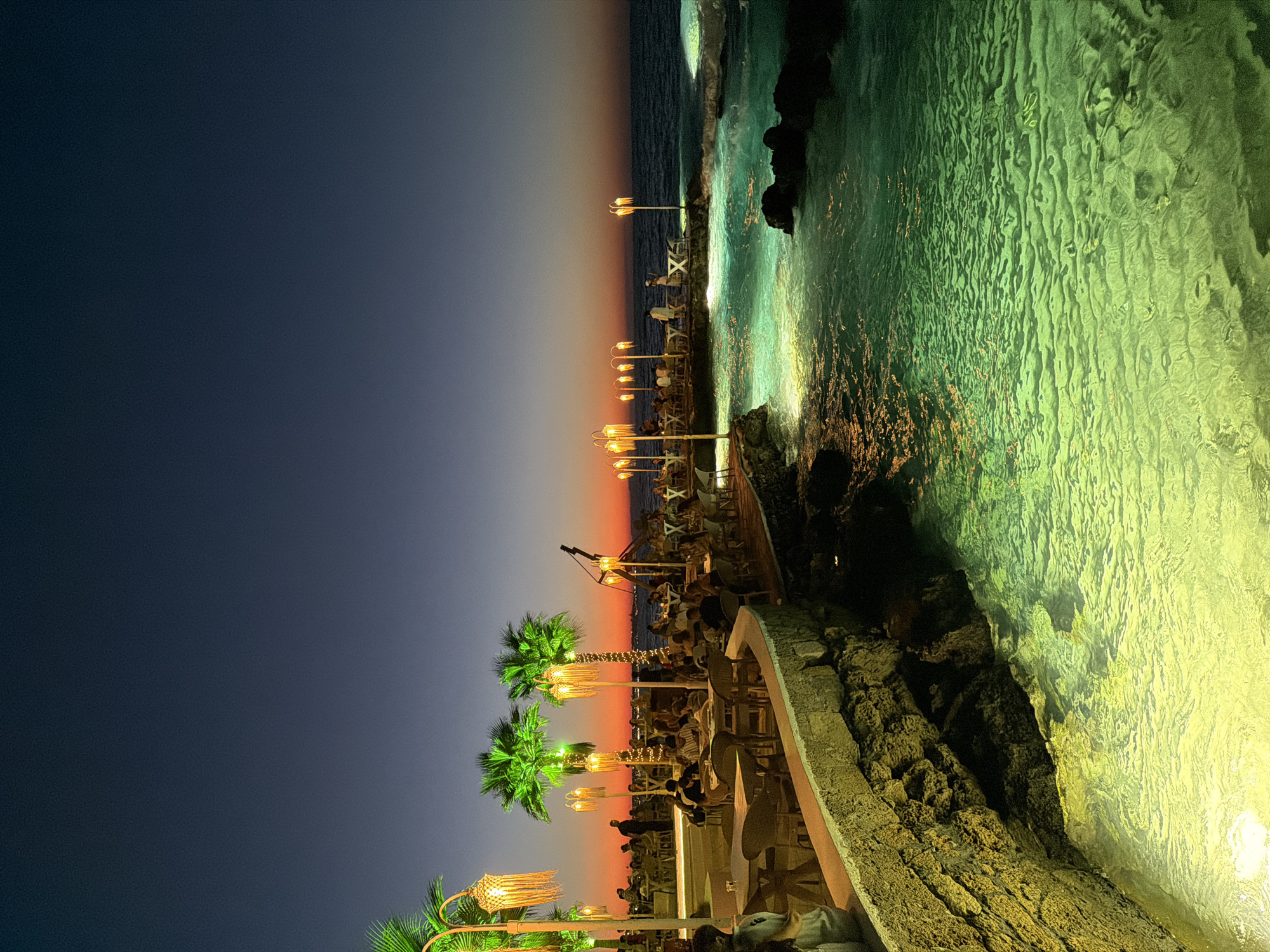 The image depicts a seaside dining establishment at dusk in Malia, Greece. The foreground features a pool of vibrant green water illuminated from below, with textured ripples and submerged sandy areas visible. Adjacent to the pool, a stone wall with rough, textured rocks forms the edge of a raised patio area. On this patio, numerous tables and chairs are set up, occupied by a crowd of people dining and socializing. Palm trees, lit with fairy lights, are interspersed amongst the seating areas, as are decorative light fixtures hanging from poles. The middle ground shows the sea with moderate waves, reflecting the warm hues of the sunset. The sky transitions from a deep blue overhead to a gradient of orange and pink near the horizon, indicating the end of daylight. Silhouettes of people and distant structures can be seen along the coastline. The overall scene suggests a lively atmosphere in a scenic, coastal location.