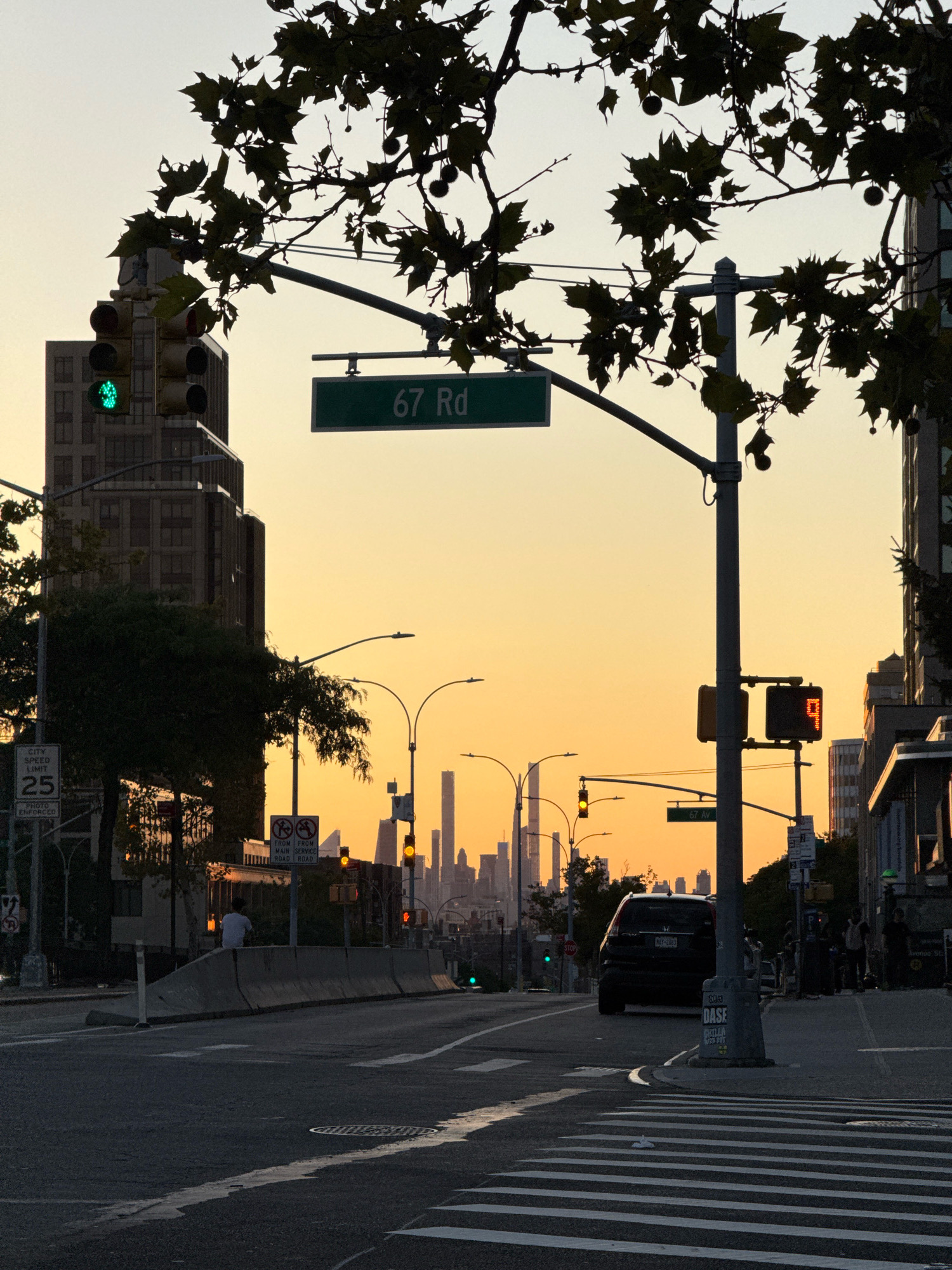 The image depicts an urban street scene at sunset, with a warm, orange glow from the sky illuminating the cityscape. The location is identified by a street sign that reads "67 Rd," indicating the intersection of 67th Road in New York City. In the foreground, a street corner with a crosswalk is visible, with a dark SUV driving away from the viewer. Traffic lights are present, one showing a green light for vehicles. A pedestrian crossing signal displays the number "9," presumably indicating the remaining seconds for pedestrians to cross. The middle ground features a concrete barrier, behind which a person is standing. Further in the background, a dense collection of skyscrapers forms the iconic New York City skyline. Silhouetted tree branches with leaves overhang the scene from the top of the frame, adding a natural element to the urban environment. The overall atmosphere is that of a quiet moment in a bustling city as day transitions into night.