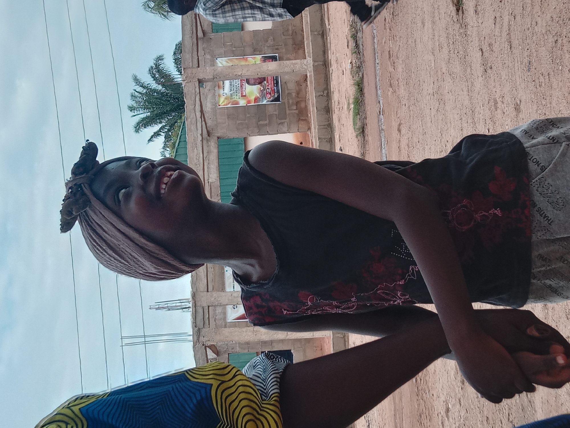The image shows a young woman in Sabon Tasha, Nigeria, looking upward and smiling. She is wearing a headwrap and a dark top with red floral accents. Her arms are crossed in front of her. In the background is a partially constructed building with exposed rebar and a palm tree. A sign is attached to the building. A partially visible individual in a patterned shirt is located at the top right.