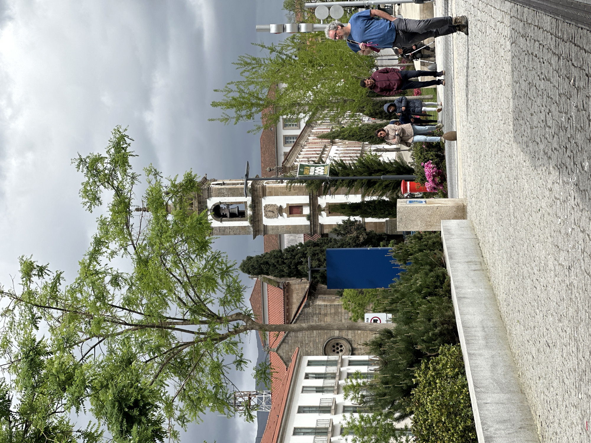 The image depicts a street scene in Vila Real, Portugal, under a cloudy sky. In the foreground, a large tree with green foliage dominates the left side. A stone wall runs parallel to the street on the right. A group of people walks along the street, including a man with a blue shirt and gray hair at the front. Behind him are three more people. In the background, architectural details include a church with a visible bell tower and white buildings with red-tiled roofs. Vegetation, including trees and shrubs, is interspersed throughout the scene. A blue sign is located in the midground. The cobblestone pavement is also visible.
