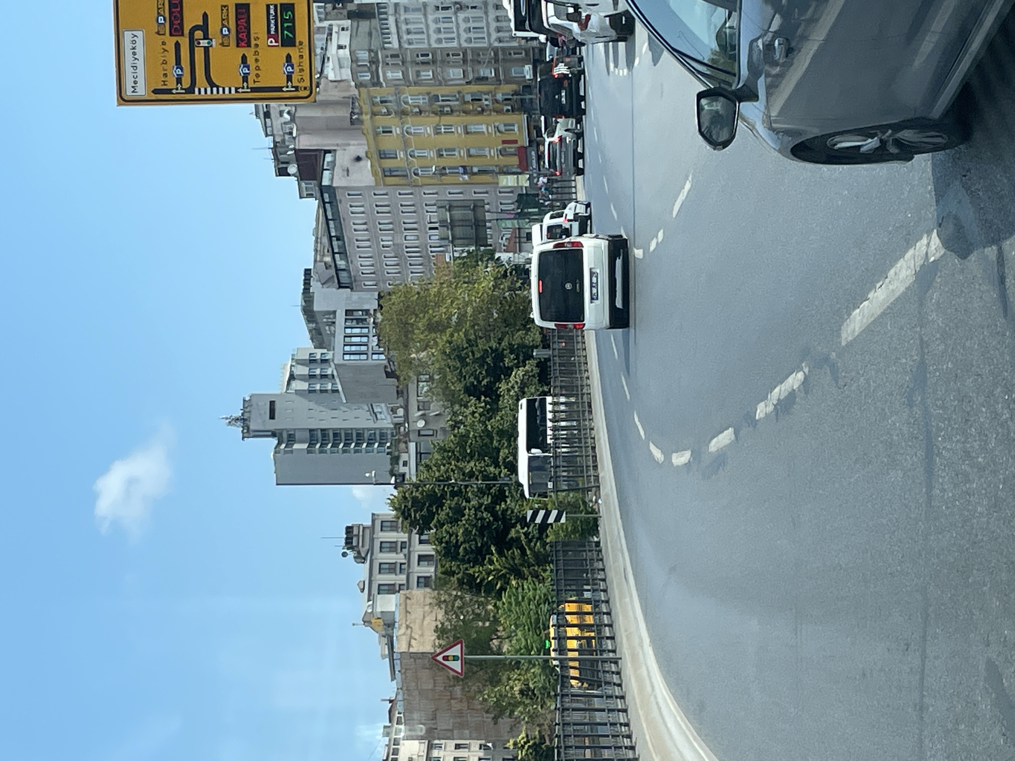 The image captures a street scene in Beyoğlu, Turkey, on a clear day with a few scattered clouds. A yellow directional sign with multiple destinations and parking symbols is prominently visible on the right side of the frame, indicating "Mecidiyeköy," "Harbiye," "KAPALI," "Tepebaşı," and "Sishane," along with route numbers and "PARK" notations. The road ahead curves to the left and is marked with dashed white lines. Several vehicles are on the road, including a white minivan in the foreground with its brake lights illuminated, a white bus further back, and other cars. A yellow taxi with a green light on top is visible behind a metal railing on the left. In the background, a collection of urban buildings, ranging in height and architectural style, line the street. Trees and greenery are interspersed among the buildings. The overall impression is of a busy city street with traffic and urban infrastructure.