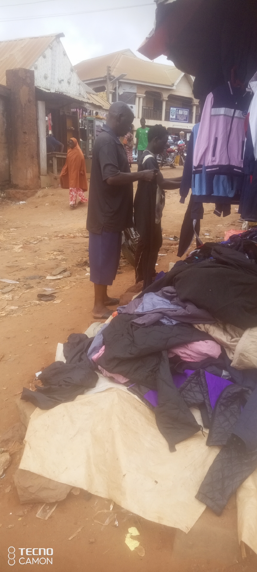 In Ankpa, Nigeria, a market scene unfolds with a focus on clothing vendors. Two individuals, an adult male and a younger male, are engaged in examining a pile of clothing. The adult male, wearing a dark t-shirt and blue shorts, stands to the left, while the younger male, in a black and white t-shirt and black pants, is to his right. They appear to be interacting with items from the pile, possibly a transaction or a selection process.
Behind them, other people are visible, including a woman in an orange headscarf and patterned skirt walking away from the camera. In the background, a building with signage that reads "S. MAN TECH" suggests a commercial area. The environment is a dirt ground, characteristic of an outdoor market, with various items of clothing displayed on tarps and possibly on a stall. The weather appears overcast. The overall impression is a busy marketplace environment.