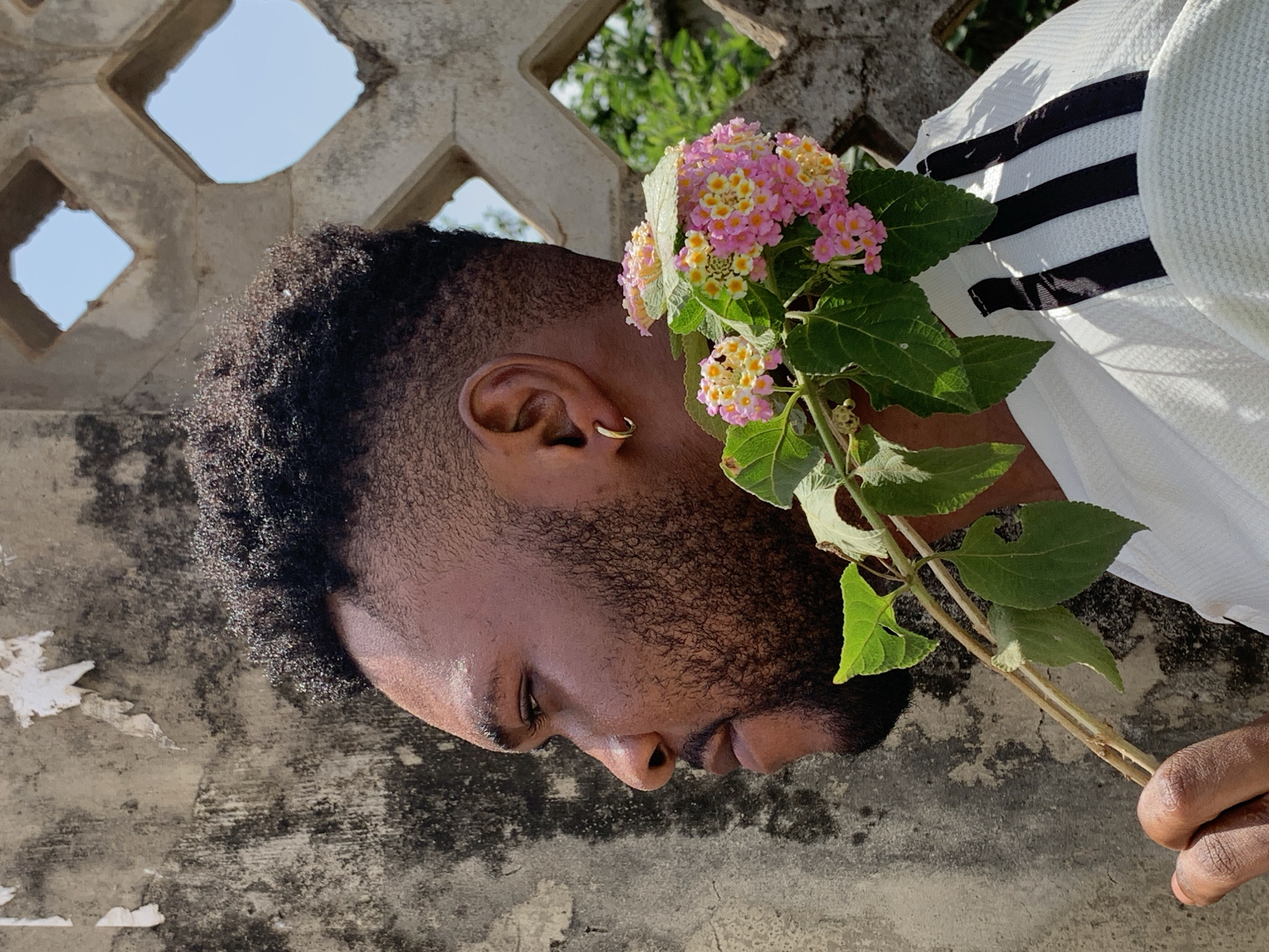 A man with a dark complexion, close-cropped black hair, and a gold hoop earring is holding a sprig of pink and yellow lantana flowers. He is wearing a white shirt with black stripes on the shoulder. He is standing in front of a concrete wall with square openings, with greenery visible through the openings. Based on the prompt, the location is Ikorodu, Nigeria.