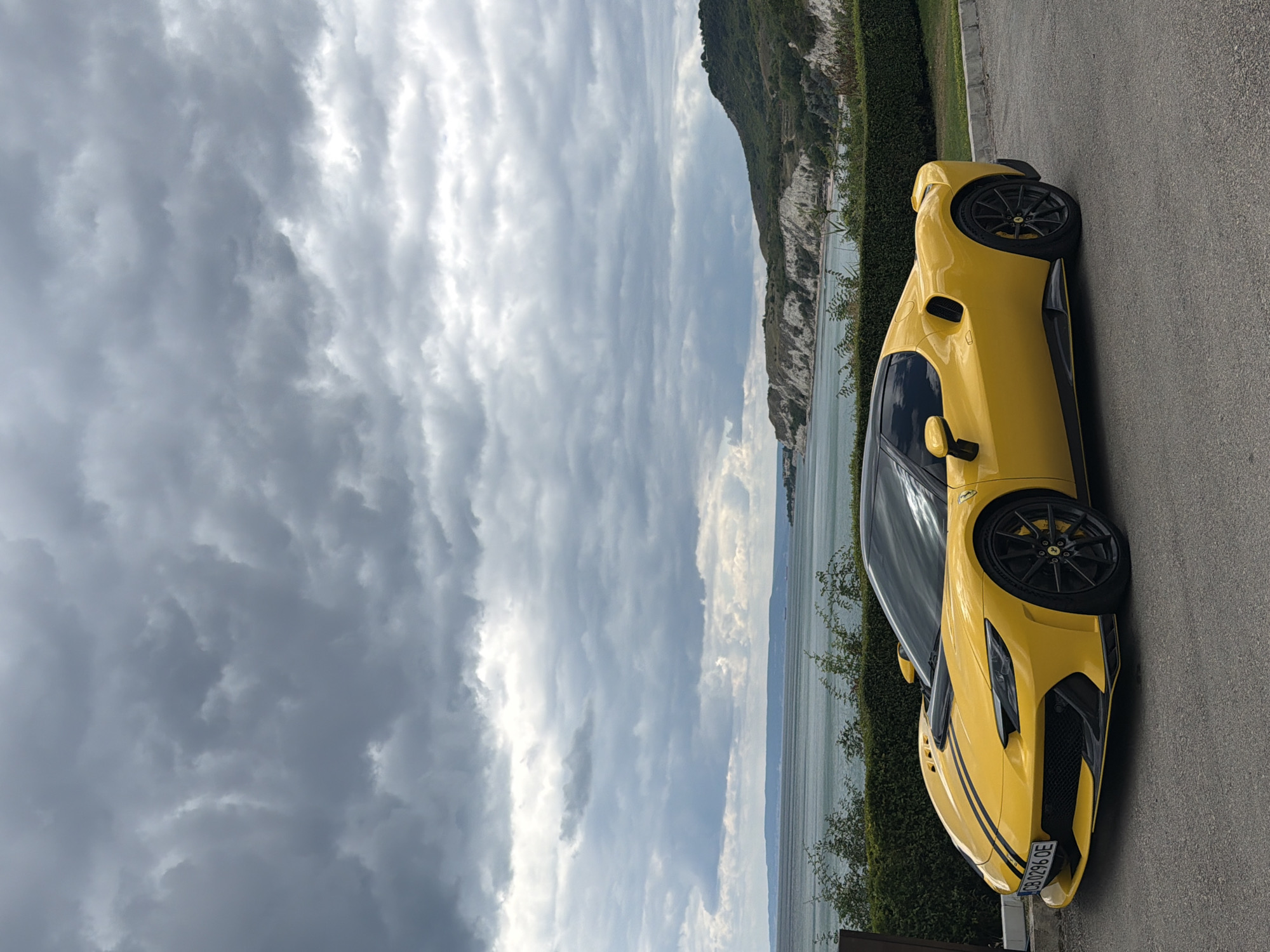 A yellow Ferrari sports car is parked on an asphalt surface in front of a neatly trimmed green hedge. Beyond the hedge, a calm sea is visible under a dramatic, cloudy sky. The coastline to the right features a green, hilly landscape with white, rocky cliffs. The car's front license plate reads "CB 0296 OE". The overall scene suggests an outdoor location near a body of water, possibly during overcast weather conditions.
