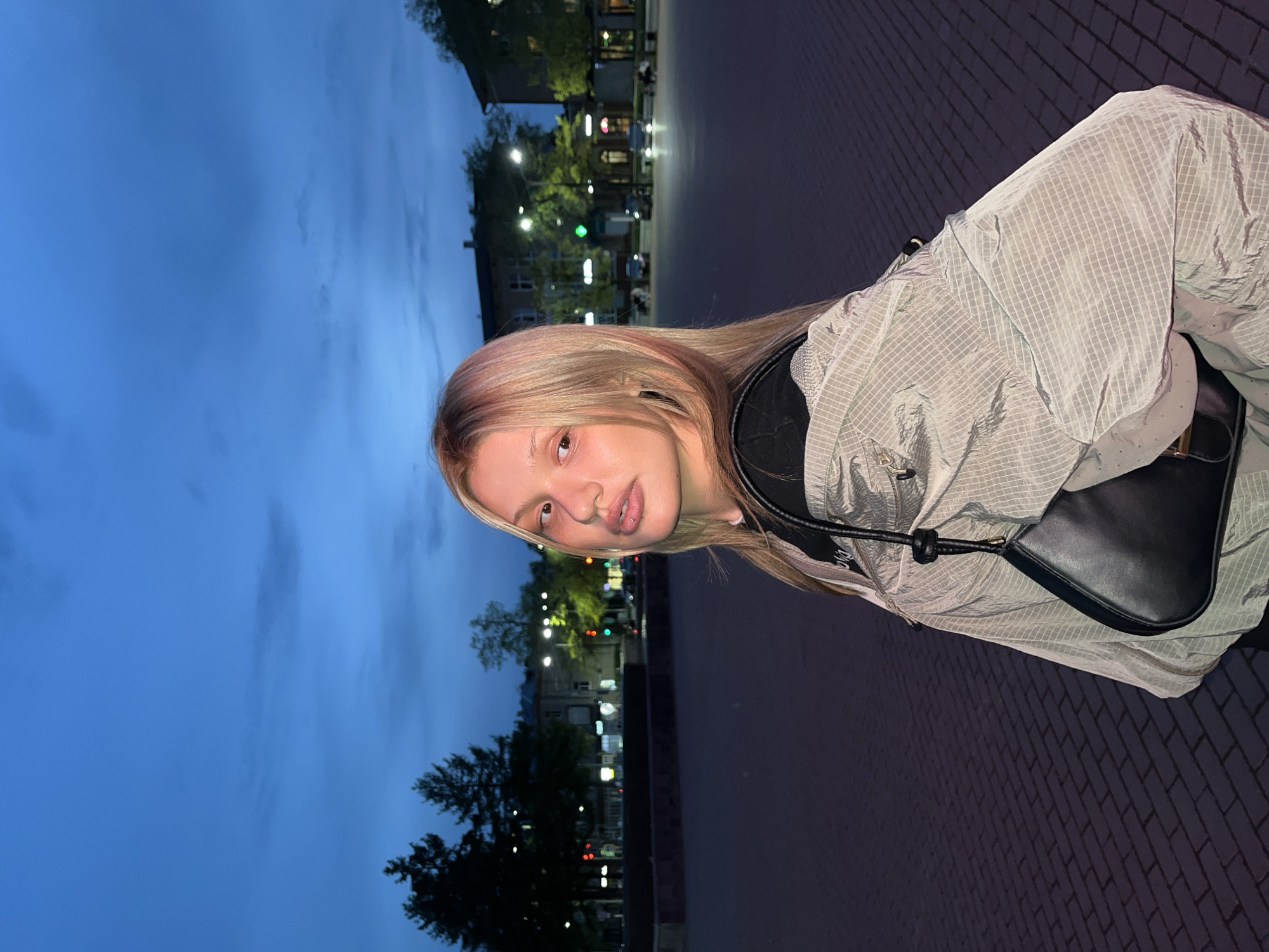 A young woman with blonde hair is standing outdoors at dusk. She is wearing a gray grid-patterned jacket, a black shirt, and a black purse slung over her shoulder. The background shows a city street with buildings, trees, and streetlights. The location is Chaikovsky, Russia.
