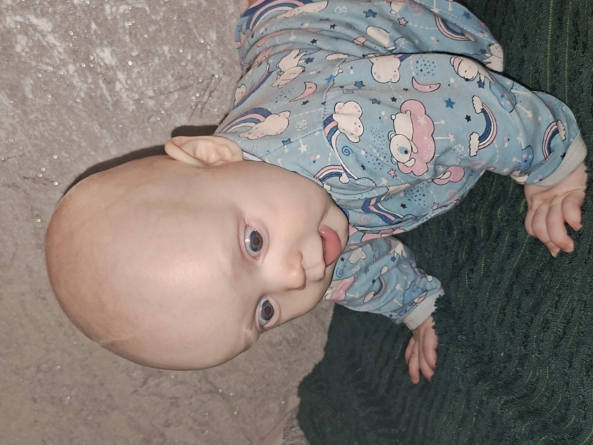 A baby lies on a gray surface on its left and a green shag rug on its right, wearing a light blue pajama top with a cartoon pattern of clouds, stars, rainbows, and teddy bears. The baby's head is bald with blue eyes, and the tongue is slightly protruding. The location is unknown.