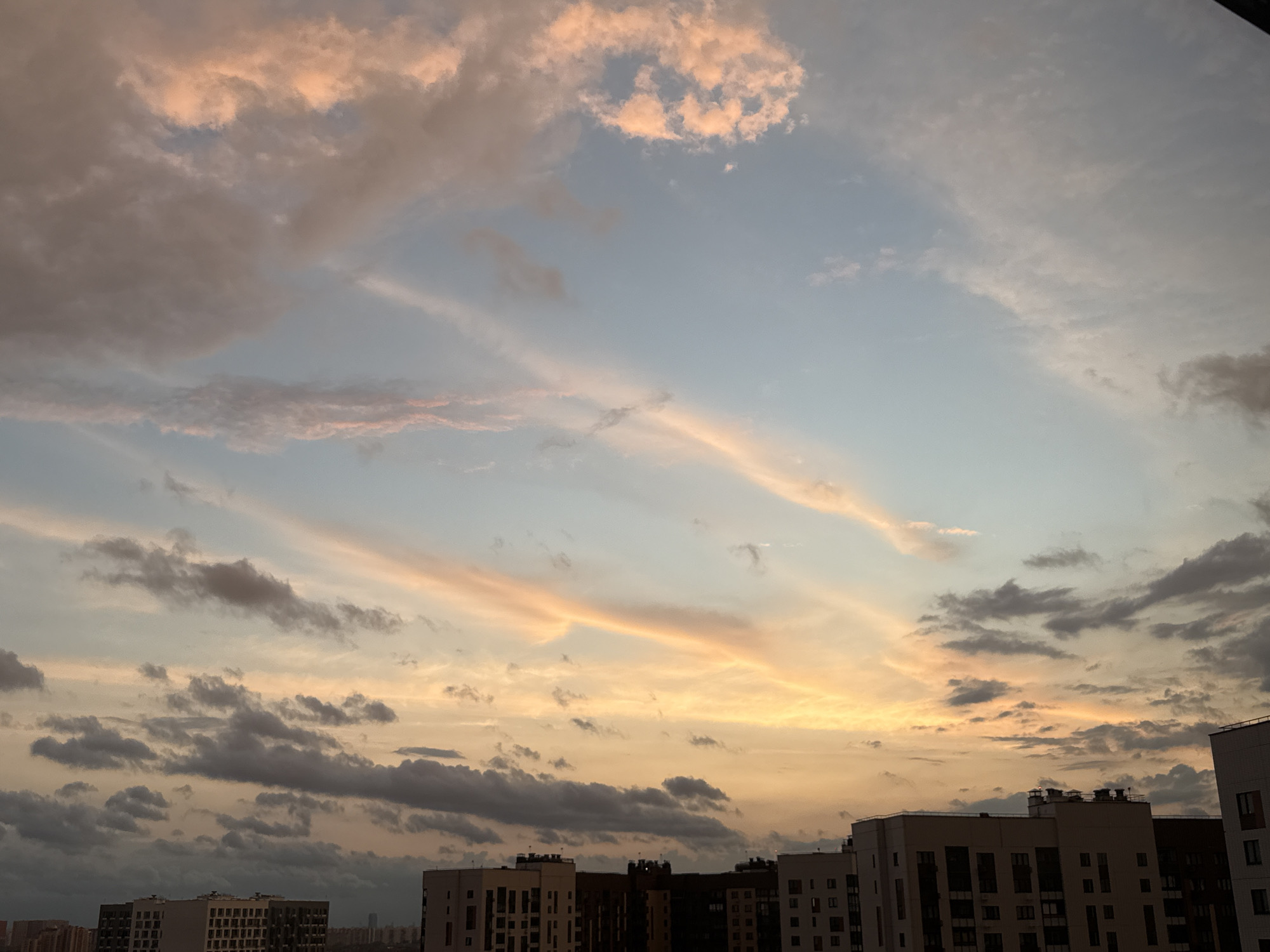 The image shows a wide shot of the sky above the city of Lyubertsy, Russia, during what appears to be sunset or sunrise. The sky is a gradient of light blue at the top, transitioning to pale yellow and orange hues near the horizon, indicating twilight. Wispy, elongated clouds are scattered across the sky, tinged with pink and orange from the low sun. Darker, more compact clouds are visible in the lower part of the sky, some appearing to be on the horizon line.
Below the sky, the tops of several multi-story residential buildings are visible. These buildings are modern in design, with numerous windows and balconies. The architecture suggests an urban environment. The overall impression is one of a city skyline set against a dramatic sky. There are no people or overt actions depicted; the scene focuses on the atmospheric conditions and the urban landscape.