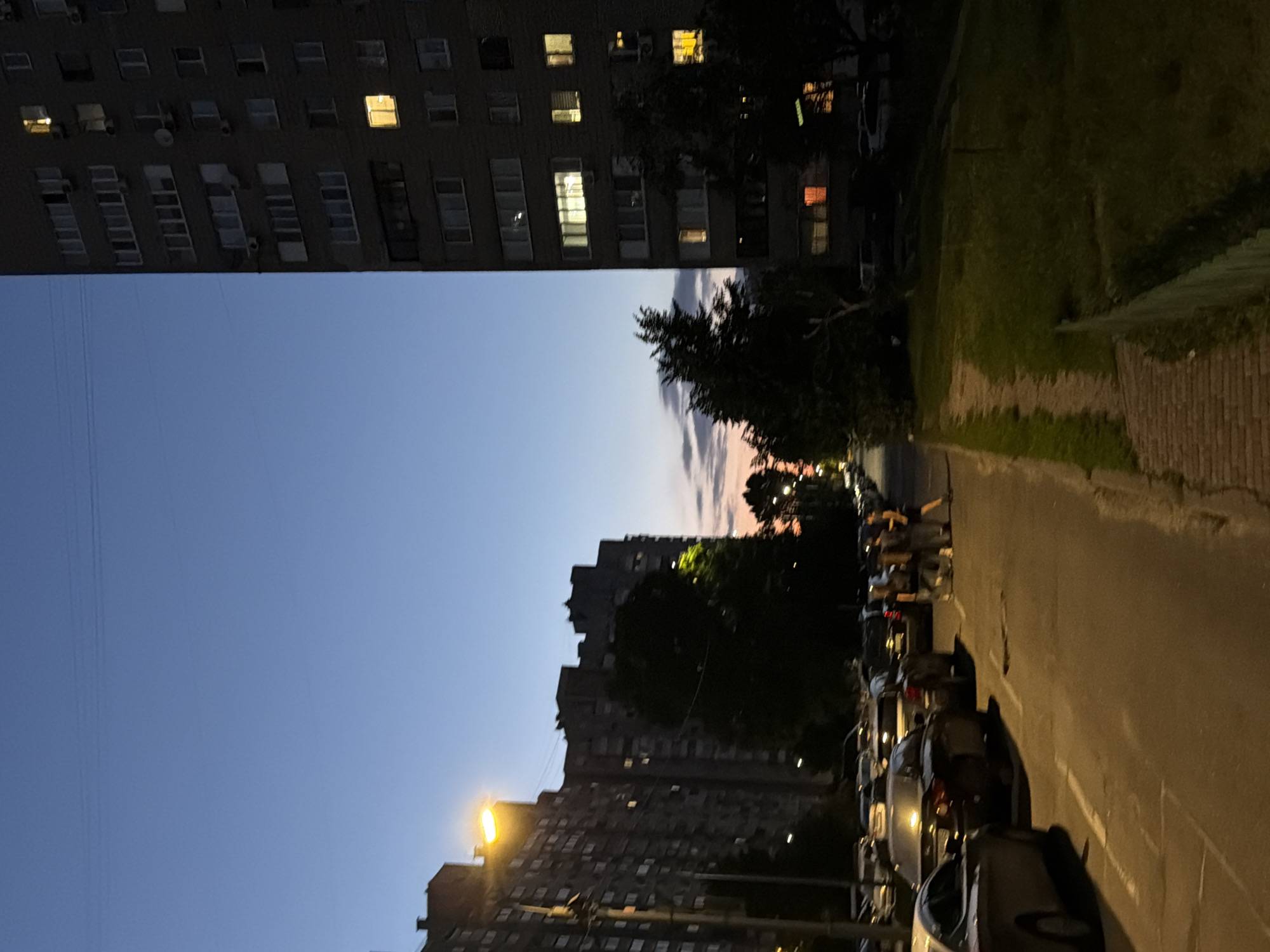 The image depicts a street scene in Kyiv, Ukraine, during dusk. The sky is a gradient of deep blue to a lighter hue near the horizon, with faint clouds visible. Two multi-story apartment buildings dominate the scene. The building on the right is tall and dark, with numerous windows, some illuminated by warm interior lights. The building on the left is also a large apartment block, with a street lamp casting a bright yellow light on its side.
In the foreground, several cars are parked along the street and on the side of the road. A group of four people, two men and two women, are standing on the sidewalk, appearing to be engaged in conversation. The street is paved, with some cracks and imperfections visible. To the right of the street, there is a grassy area with a low green fence. Trees are scattered throughout the scene, some with dark foliage and others silhouetted against the twilight sky. The overall impression is of a residential area in an urban environment as day transitions to night.