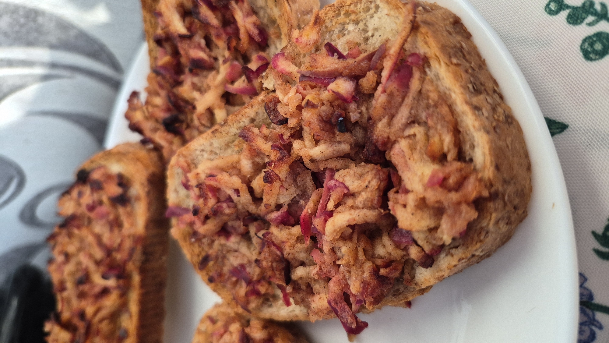 The image displays a close-up, eye-level shot of a white plate holding several slices of toasted bread topped with what appears to be a grated apple and cinnamon mixture, possibly baked or caramelized. The bread slices are multigrain and appear toasted to a light brown. The topping is a reddish-brown color with visible shreds of apple and spices, some pieces slightly charred. The plate is positioned on a tablecloth with a subtle floral pattern. The background is out of focus, showing a textured surface and a hint of a chair or furniture. The scene appears to be indoors, possibly in a domestic setting. The location is not specified in the image.
