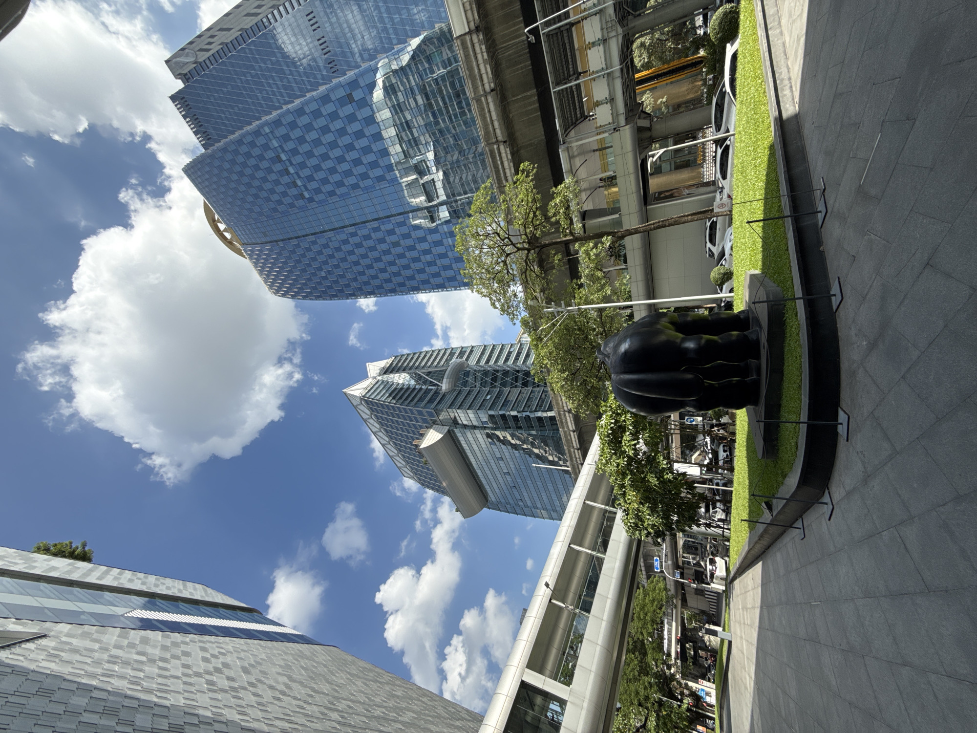 This is a low-angle, wide-shot photograph taken in Bangkok, Thailand, likely in the vicinity of the BTS Skytrain elevated track and surrounding modern architecture. In the foreground, a dark, matte sculpture, possibly of an animal's hindquarters, is positioned on a manicured patch of grass. The sculpture is low to the ground and its texture appears rough. Beyond the sculpture and grass, a paved walkway is visible, leading towards a street with vehicles.
The mid-ground is dominated by elevated structures, including a silver, enclosed pedestrian walkway that bridges across the scene. Below this walkway, an elevated train track, presumably part of the BTS Skytrain system, runs horizontally. Green trees with dense foliage are interspersed between these structures, adding a natural element to the urban landscape.
In the background, towering modern skyscrapers with reflective glass facades are prominent. One building on the right features a checkered pattern of blue and darker panes and is crowned with a distinctive circular, gold-colored structure. Another building on the left has a more textured, gray facade and sharp, angular architectural features. The sky is a bright blue with scattered, fluffy white clouds. The overall impression is one of a bustling, contemporary urban environment with a blend of sleek architecture, public transportation infrastructure, and some pockets of greenery.