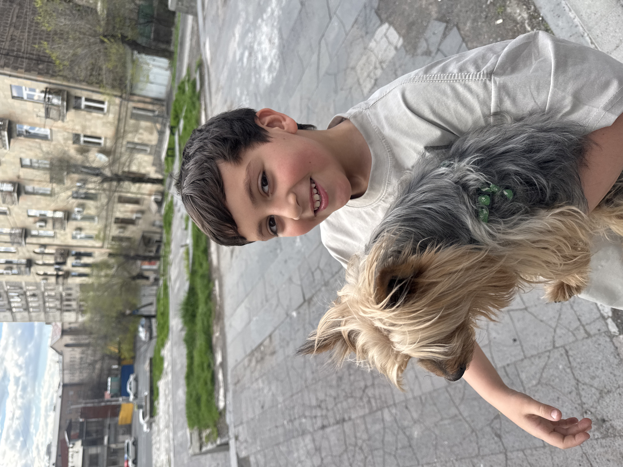 A young boy is holding a Yorkshire Terrier in Yerevan, Armenia. The boy has dark hair and a light complexion. He is wearing a light-colored t-shirt. The dog has gray and tan fur and a green hair accessory. They are standing on a gray brick or concrete surface. In the background, there are buildings characteristic of Yerevan architecture, street, and greenery. The picture has been rotated and needs to be viewed sideways.
