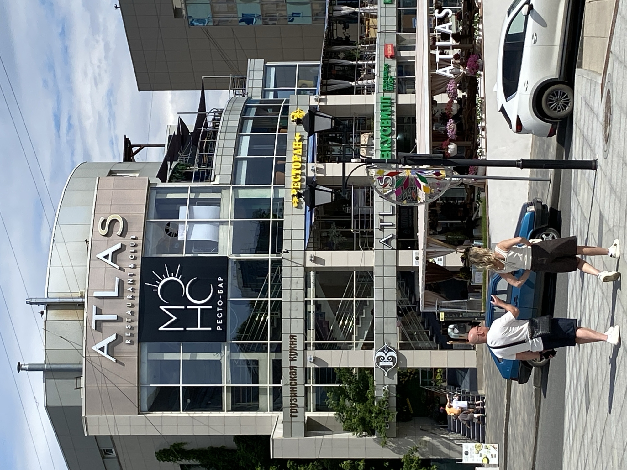 This media file captures an outdoor scene on a sunny day, with a modern building as the primary subject. The building features a glass facade and a prominent sign that reads "ATLAS RESTAURANT & CLUB" with the "MHC" logo and "РЕСТО-БАР" below it. Another sign in green letters says "ВКУСОВИЛ!". A sign in Russian on a lower level says "грузинская кухня" (Georgian cuisine). There are several people visible. In the foreground, two individuals are standing on the sidewalk, one with a phone in hand. Further back, a white car is parked on the street, and a blue car is also present. People can be seen walking and sitting on outdoor seating associated with the establishment. The overall impression is of a bustling urban environment with businesses open and people enjoying the day. The lighting suggests it is daytime, likely midday or afternoon.