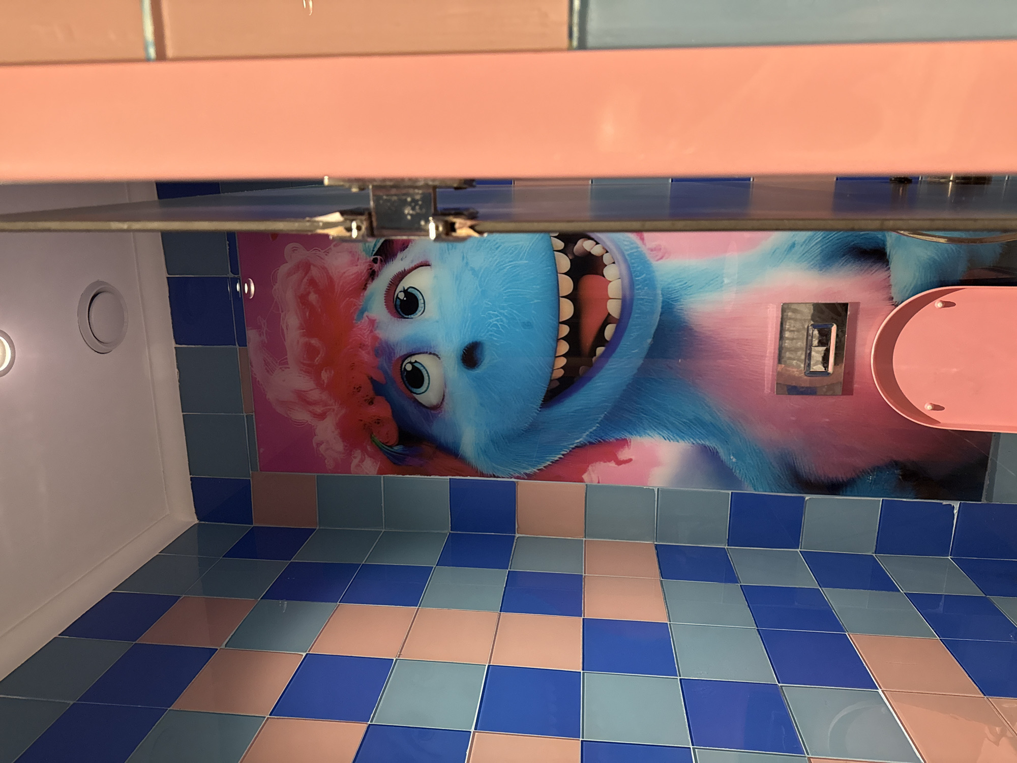 The image shows a close-up view inside a brightly colored stall, likely a restroom, in Bodrum, Turkey. The walls are tiled in a checkered pattern of blue and light pink/peach squares. On the back wall, a large decal features a cartoon character with blue fur, wide blue eyes, and a bright pink, fluffy hairdo. The character has a large, open mouth revealing many teeth and a pink tongue. In the foreground, to the right, a pale pink wall or door is partially visible, with a vertical seam and metal fixtures suggesting it might be a door or partition. A pink toilet seat is visible at the bottom of the image. The overall impression is one of a playful and vibrant aesthetic.