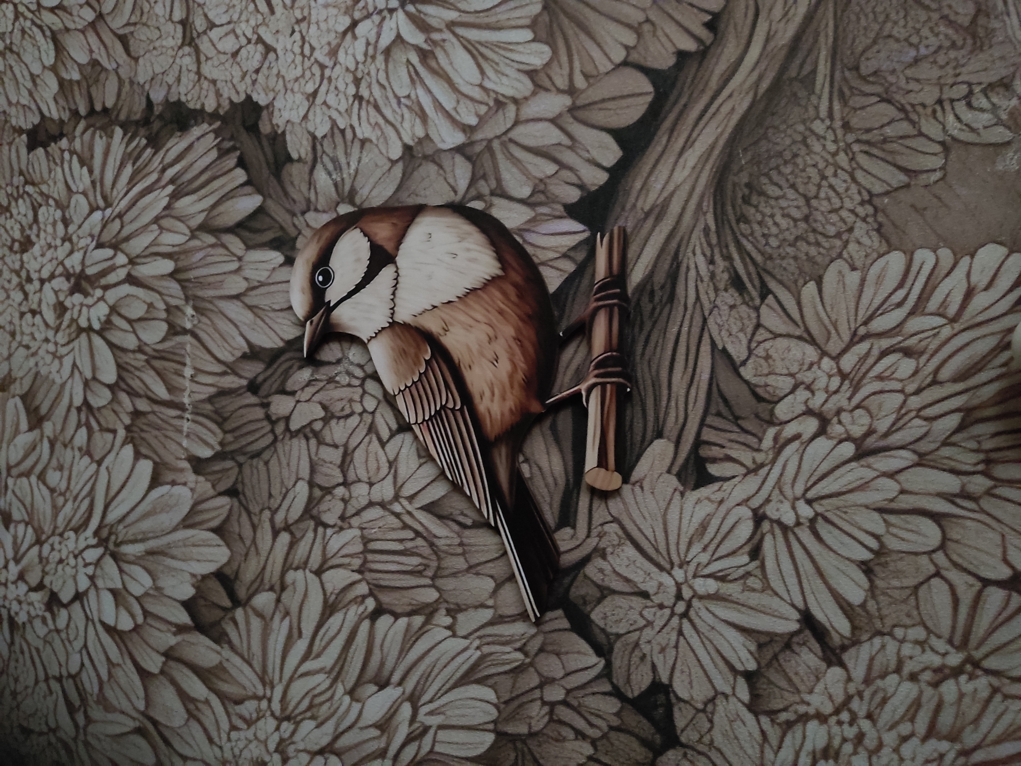 The image is a close-up, vertical shot of a sepia-toned painting or illustration of a bird perched on a branch. The bird, depicted in realistic detail with brown, white, and black plumage, faces to the left, with its head slightly tilted. It has a distinctive white stripe above its eye. The bird is perched on a horizontal wooden branch that extends diagonally from the lower left to the middle right of the frame. The background is filled with a dense pattern of stylized, feathery floral elements, also in sepia tones, creating a textured and somewhat abstract backdrop. The lighting appears even, highlighting the details of the bird and the textured background. There is no identifiable location in the city of Noginsk, Russia, within the image.