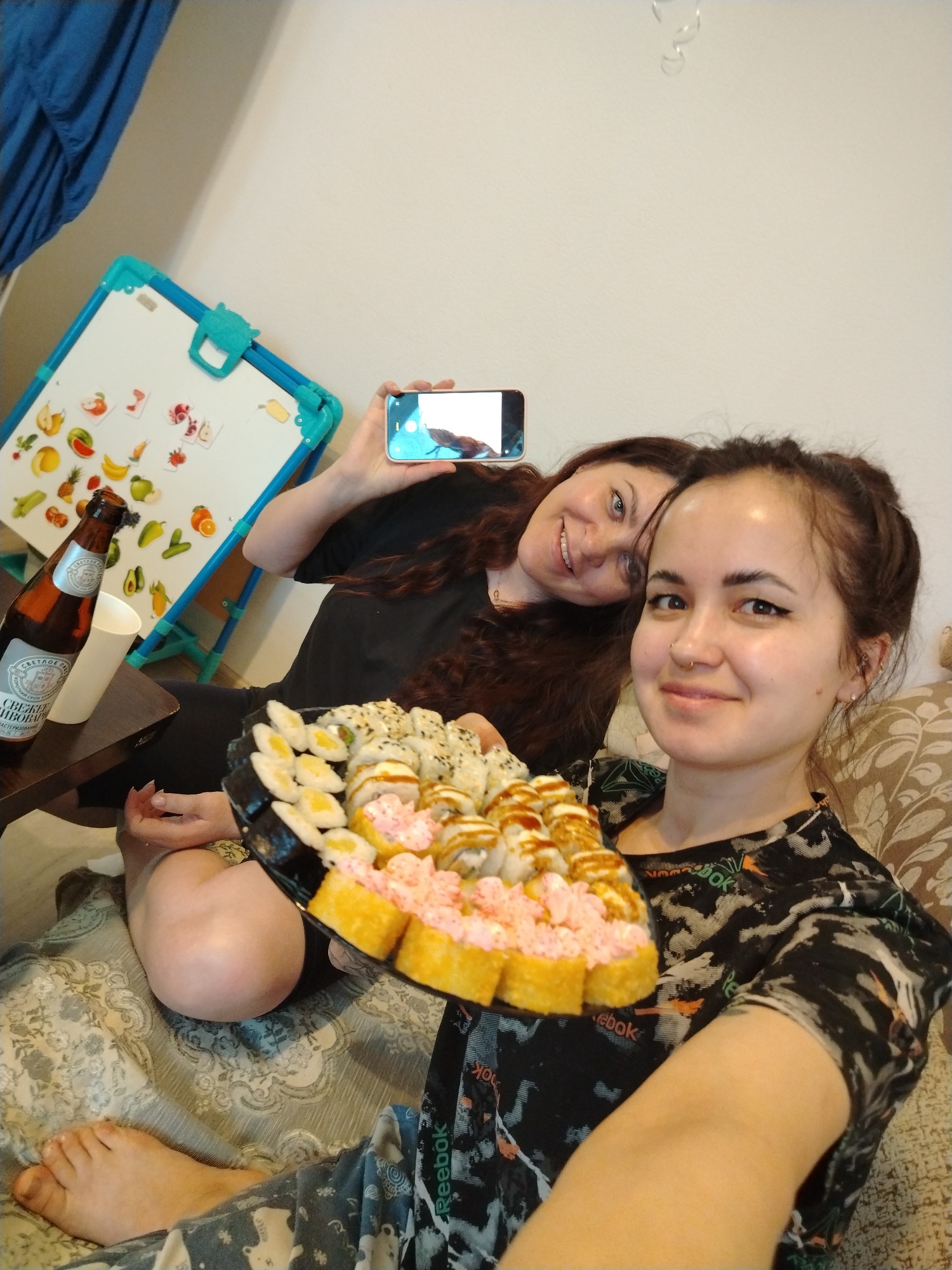 Two women are in an indoor setting, possibly a home, based on the presence of a couch, a small table, and a child's whiteboard with food magnets. The woman in the foreground is holding a large tray of sushi, while the woman in the background is holding a smartphone, presumably taking a photo or video. A bottle of beer and a white cup are on the table. The setting appears casual and relaxed.