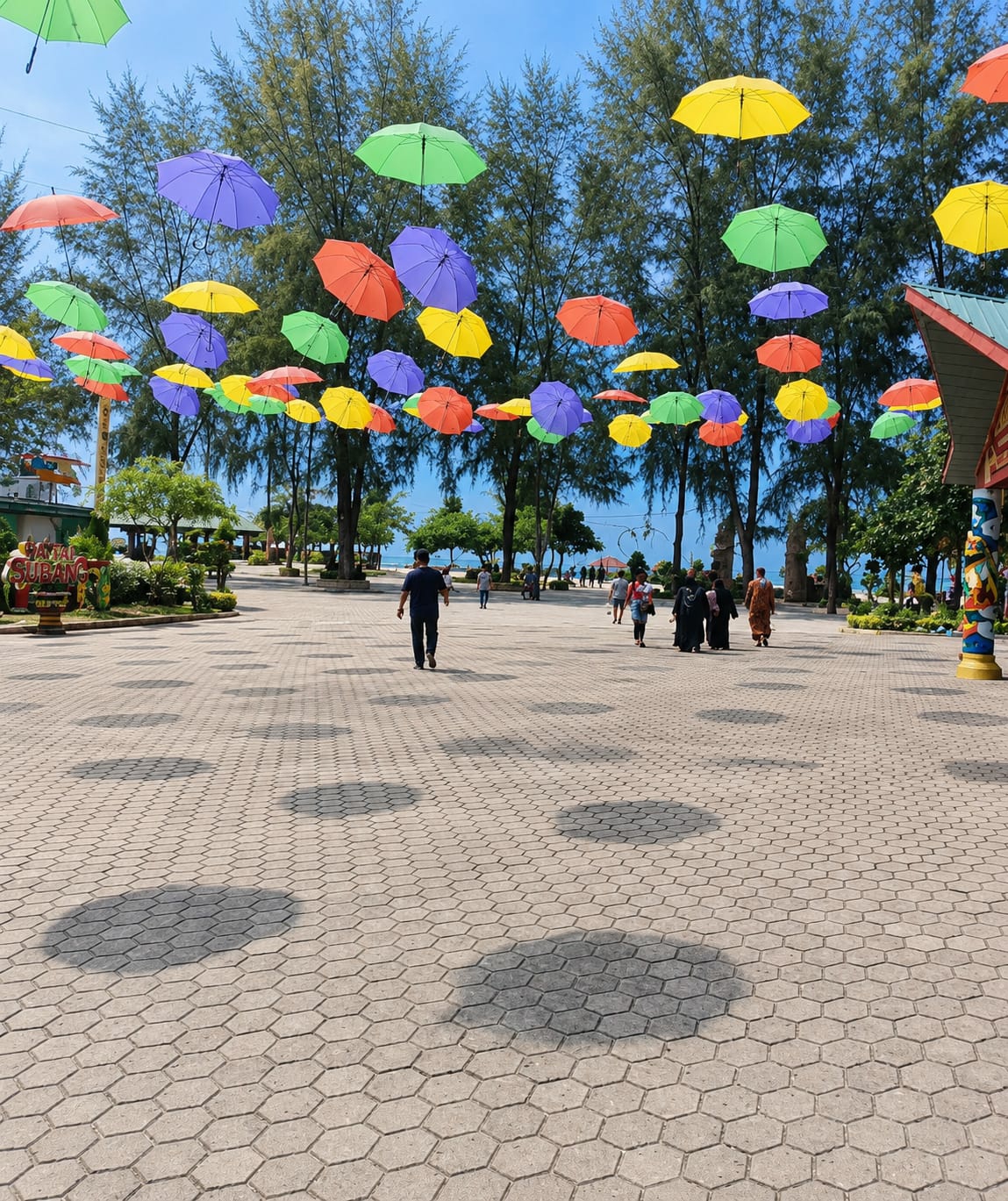 The image depicts an outdoor plaza in Kota Pari, Indonesia, adorned with numerous colorful umbrellas suspended in the air. The plaza features a hexagonal patterned pavement with circular shadows cast by the umbrellas. Several individuals are walking across the plaza. In the background, lush green trees line the walkway, and beyond them, a body of water is visible, likely the sea, with a clear blue sky above. The overall atmosphere appears to be a public recreational area, possibly a park or a promenade, decorated for a festive or artistic display.