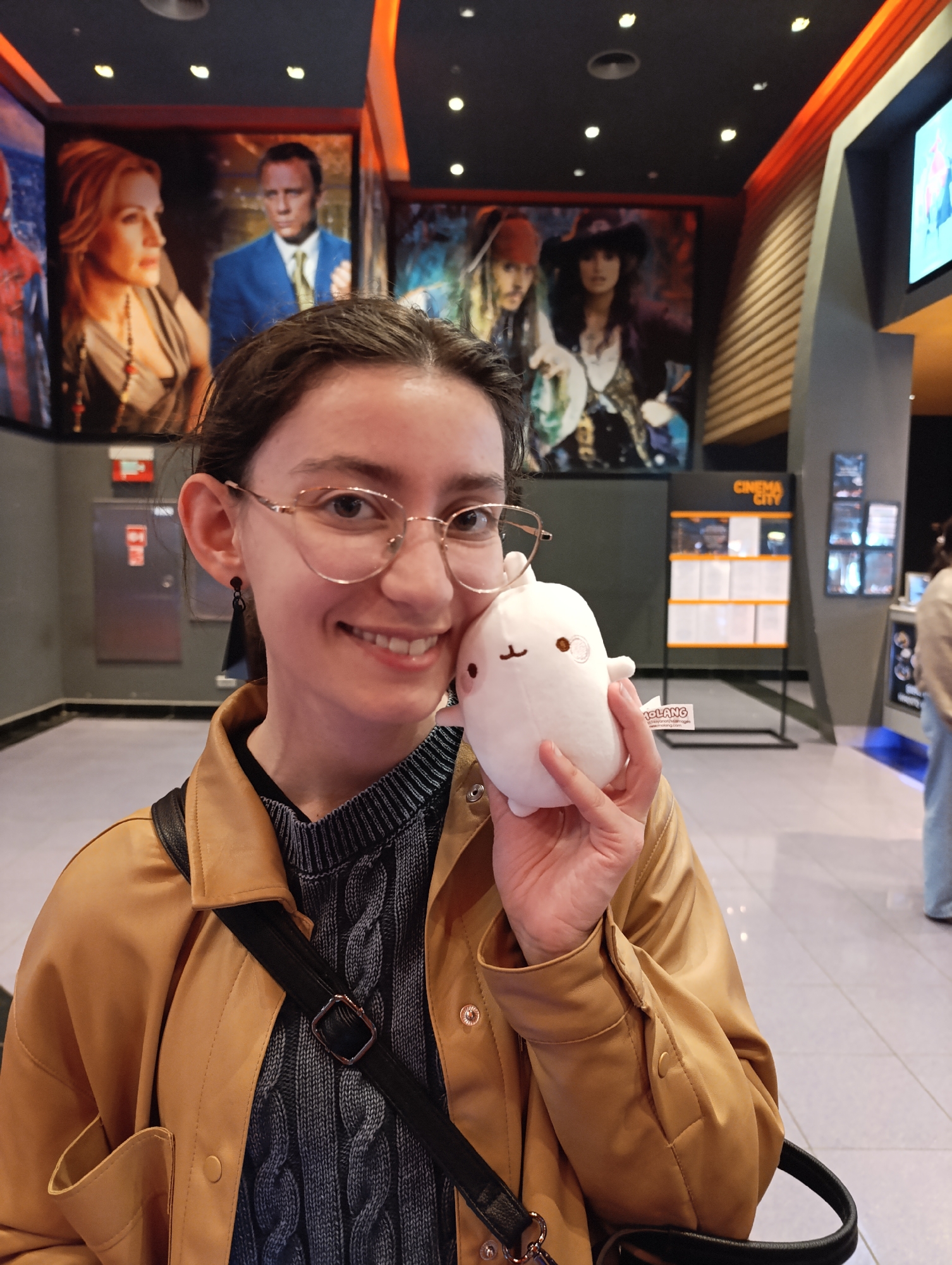 A young woman with a joyful smile poses inside a modern cinema lobby. She wears a light tan leather-look jacket over a dark blue and grey cable-knit sweater, with gold-rimmed glasses and dark dangling earrings. In her left hand, she holds a small, white, round plush toy with the word "MOLANG" visible on its tag.
The background features large movie posters: a partial view of Spider-Man on the far left, and more prominently, Daniel Craig in a blue suit beside a blonde-haired woman (likely from a James Bond film) and Johnny Depp as Captain Jack Sparrow alongside Penélope Cruz (from *Pirates of the Caribbean*). To the woman's right, a tall black and orange display board clearly shows the text "CINEMA CITY" at the top, with informational panels below it.
The interior is well-lit with numerous recessed ceiling lights and orange accent lighting along a wall feature on the right. The floor is light-colored and reflective. A red fire extinguisher cabinet is visible on the wall behind the woman to the left. The scene depicts a casual moment in a bustling cinema, likely in Sofia, Bulgaria.