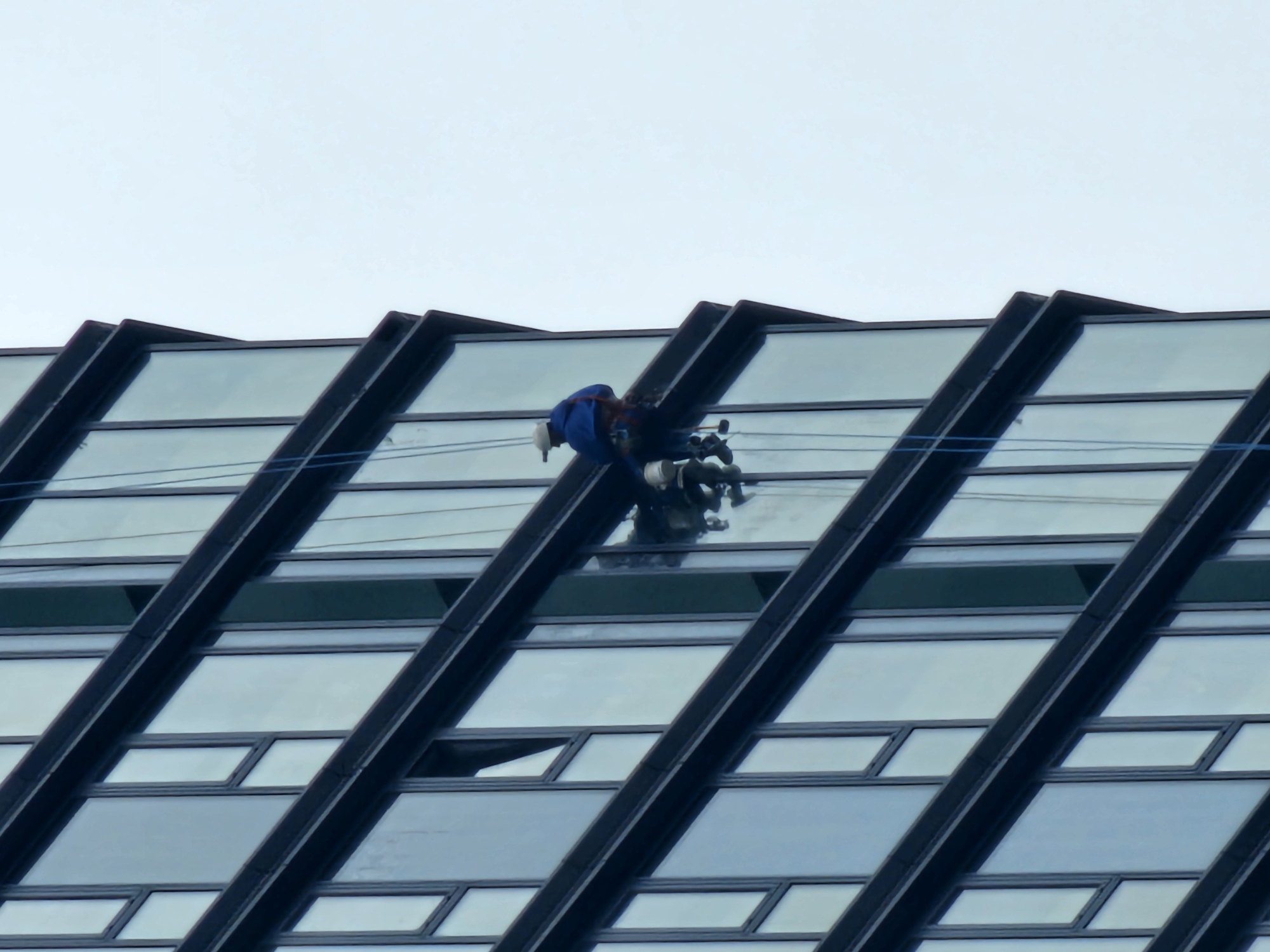 A window washer is suspended from the roof of a tall, modern building in Moscow, Russia. The building's exterior features a repeating pattern of angled glass windows separated by dark, diagonal beams. The window washer is wearing a blue uniform and a white helmet. Ropes secure the window washer to the building. The sky is overcast.