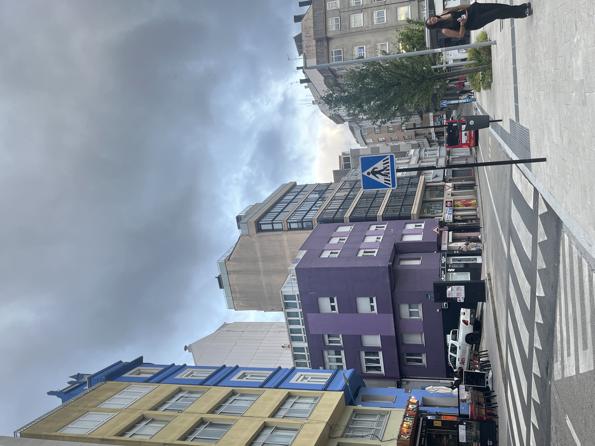 The image captures a lively urban street scene in A Coruña, Spain, under an overcast sky. The view looks along a street lined with buildings of varying architectural styles and colors.
In the foreground, on the left, a building with distinct yellow lower floors and blue upper sections features multiple windows. Adjacent to it on the right, a prominent multi-story building painted a vibrant purple stands out, also with numerous windows. Further down the street, a modern structure with a glass facade is visible, nestled among more traditional, grey apartment buildings in the background.
The street features a clearly marked zebra crossing and white road lines. A blue square sign with a white pedestrian symbol indicates the crossing, mounted on a black pole that also holds a black waste bin. A white pickup truck, with "Amstec" visible on its side, is parked on the street beside the purple building. In the distance, a red bus can be seen on the road.
On the paved sidewalk, near the top right of the frame, a woman with long dark hair, dressed in black attire, stands leaning against a pole or barrier. She is smiling broadly, looking towards the camera, and holding a small white object, possibly a phone. Other pedestrians are faintly visible further down the street. Near the yellow and blue building, an outdoor cafe area with tables, chairs, and white umbrellas suggests a bustling establishment. Visible text includes "Amstec" on the pickup truck and partial text like "LA TABERNA" and "BAR" on a menu board at the cafe. The overall atmosphere is that of a typical city day, despite the cloudy weather.