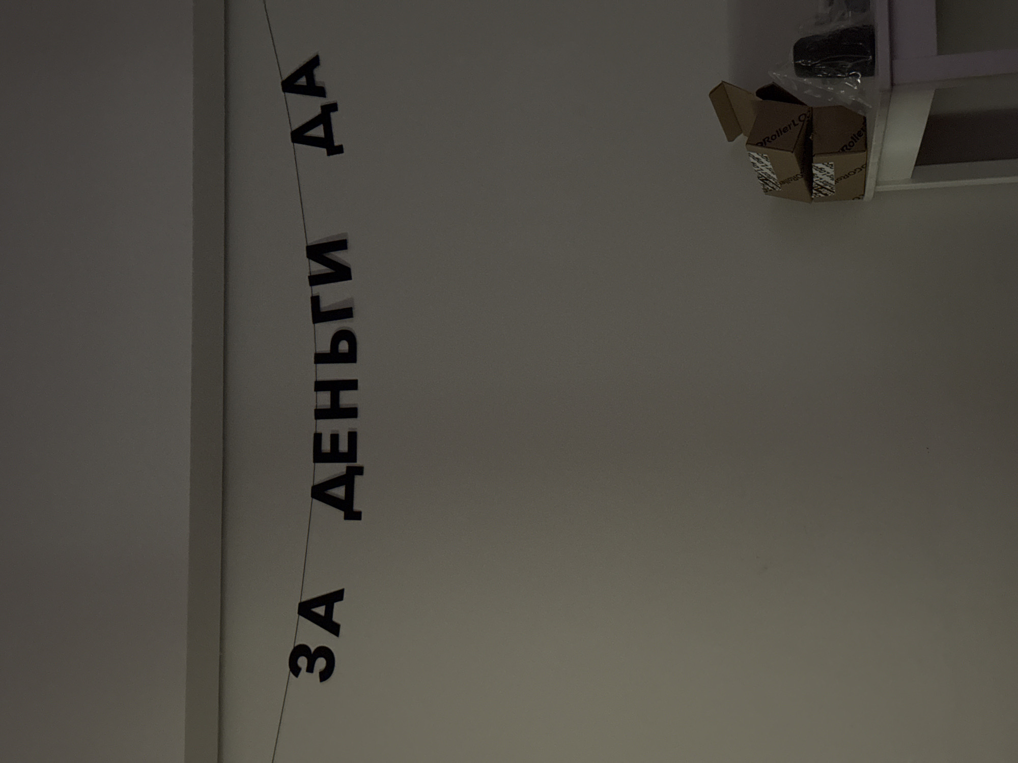 The image, oriented correctly, depicts an indoor scene with a light-colored wall serving as the main background. A decorative banner, composed of individual black capital letters strung together, hangs across the wall. The letters spell out the Cyrillic phrase "ЗА ДЕНЬГИ ДА".
In the upper right portion of the frame, a white shelf or ledge is visible, holding two small, open cardboard boxes. One of these boxes has the text "RollerLO" printed on its side, along with some clear plastic packaging materials. To the left, a subtle vertical line suggests the edge of a wall or an architectural feature.
No people are present, and no direct activity is taking place. The scene appears to be a static interior setting, possibly a room in an office or a home in Moscow, Russia. The lighting is ambient and even. The Cyrillic text "ЗА ДЕНЬГИ ДА" translates to "For money, yes," suggesting a message related to commercial activity, payment, or a humorous take on willingness to perform tasks for monetary compensation.