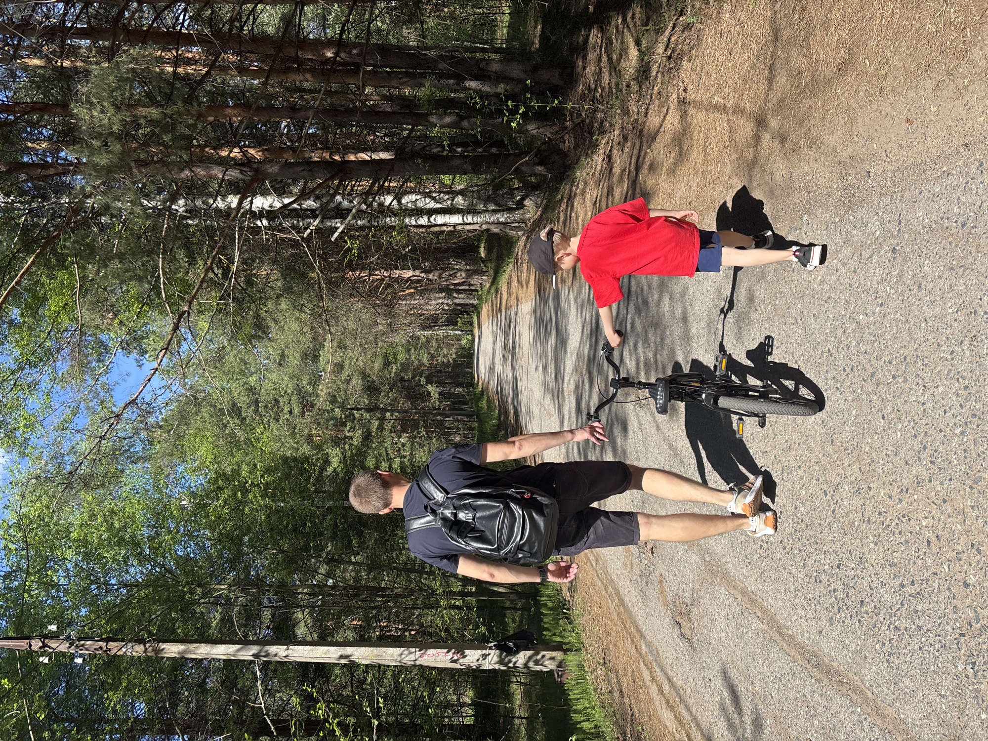 Two people, one adult male and one child, are standing on a paved road in a wooded area. The adult male is wearing a dark t-shirt, dark shorts, a black backpack, and tan shoes. The child is wearing a red t-shirt, dark shorts, a black baseball cap, and black and white shoes. A black bicycle is lying on the ground between them. The road is surrounded by trees, and overhead power lines are visible to the left side of the frame. The location is Chaikovsky, Russia.