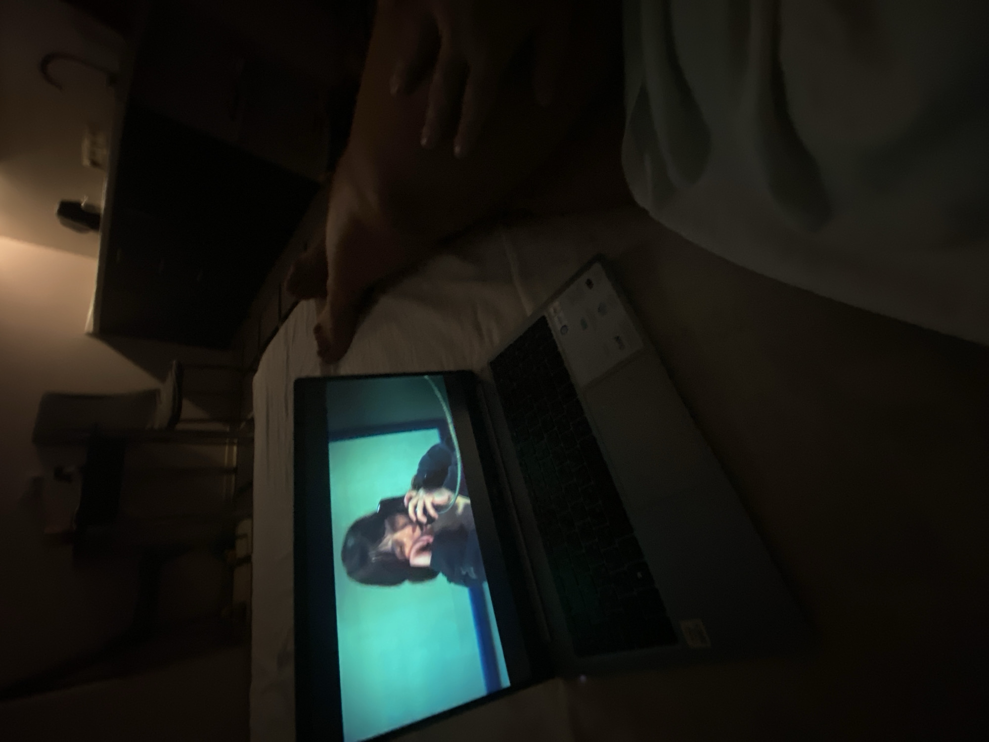 A close-up shot captures a laptop displaying a video of a person on the phone, set against a blurred background. A person's legs and hand are visible in the lower right portion of the frame, suggesting they are lying down. The overall lighting is dim, creating a sense of intimacy. The scene is not definitively identifiable as being in Sochi, Russia, or any other specific city.