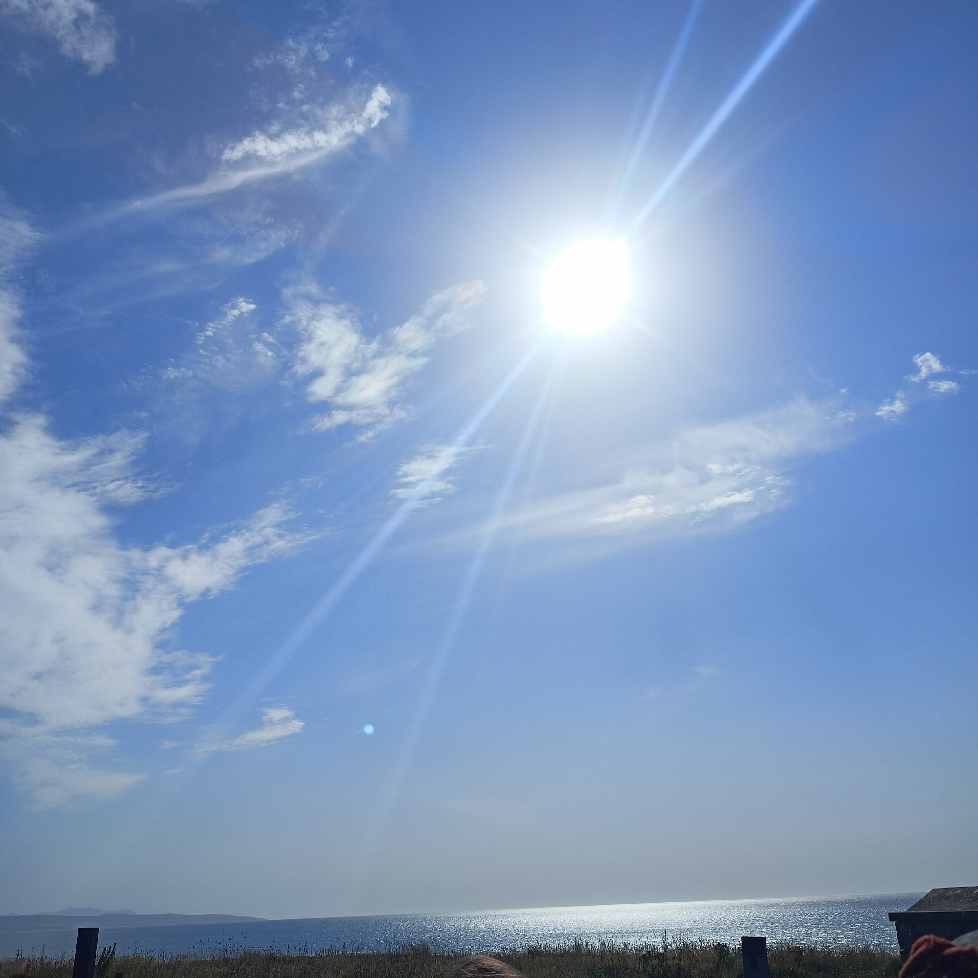 This image depicts a bright, sunny day at Ballybunnion, Ireland. The sun is high in the sky, creating a strong lens flare and illuminating the clear blue sky with scattered white clouds. In the foreground, a calm body of water, likely the Atlantic Ocean, stretches to the horizon. The shoreline is lined with dry grass and vegetation. A fence post is visible on the left, and a section of a stone building is on the right. The overall scene conveys a peaceful, natural setting by the coast.