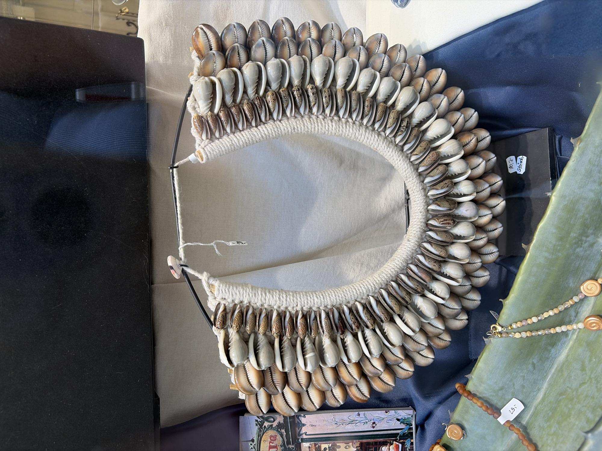 This image shows a display of necklaces in what appears to be a shop window. The primary object of focus is a large, ornate necklace made of numerous cowrie shells, arranged in multiple rows around a thick, woven rope. The shells are a mix of cream, brown, and white, with varying patterns. The necklace is suspended from a dark metal frame. To the left, partially visible, is a decorative item with images of buildings and figures, possibly a piece of artwork or a decorative box. In the foreground, resting on a broad, green, spiky leaf, are two other necklaces. One is made of small, brown, spherical beads and has a price tag attached. The other is a delicate strand of small, multi-colored beads and charms. The background consists of draped fabric in shades of beige and dark blue, and a black, reflective surface which may be part of the shop's display or a window. The overall impression is a retail setting, likely a souvenir shop or a boutique selling handcrafted jewelry. The specific location within Würzburg, Germany, cannot be determined from the visual information provided.