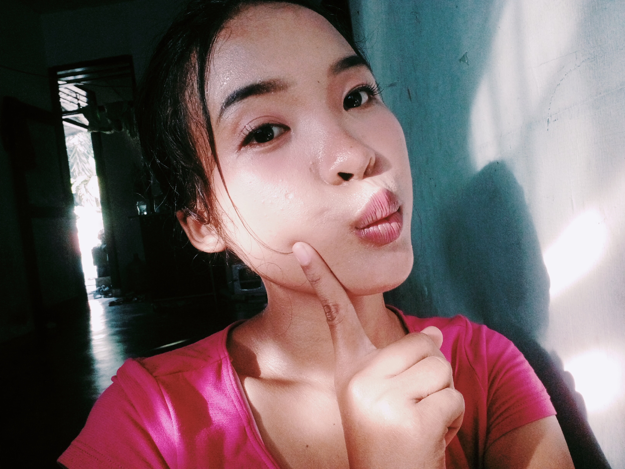 The image shows a young woman in a pink t-shirt, taking a selfie. She has her right index finger touching her chin, and her lips are pursed as if she is about to kiss. The background is a wall with light and shadows cast on it, possibly from a window or doorway. The room appears to be dimly lit, with sunlight streaming in from an opening on the left side of the image, illuminating a dark hallway or room with a reflective floor. The overall impression is that the photograph was taken indoors, likely in a residential setting in Pamesi, Indonesia, given the lighting and architectural elements suggested by the background.