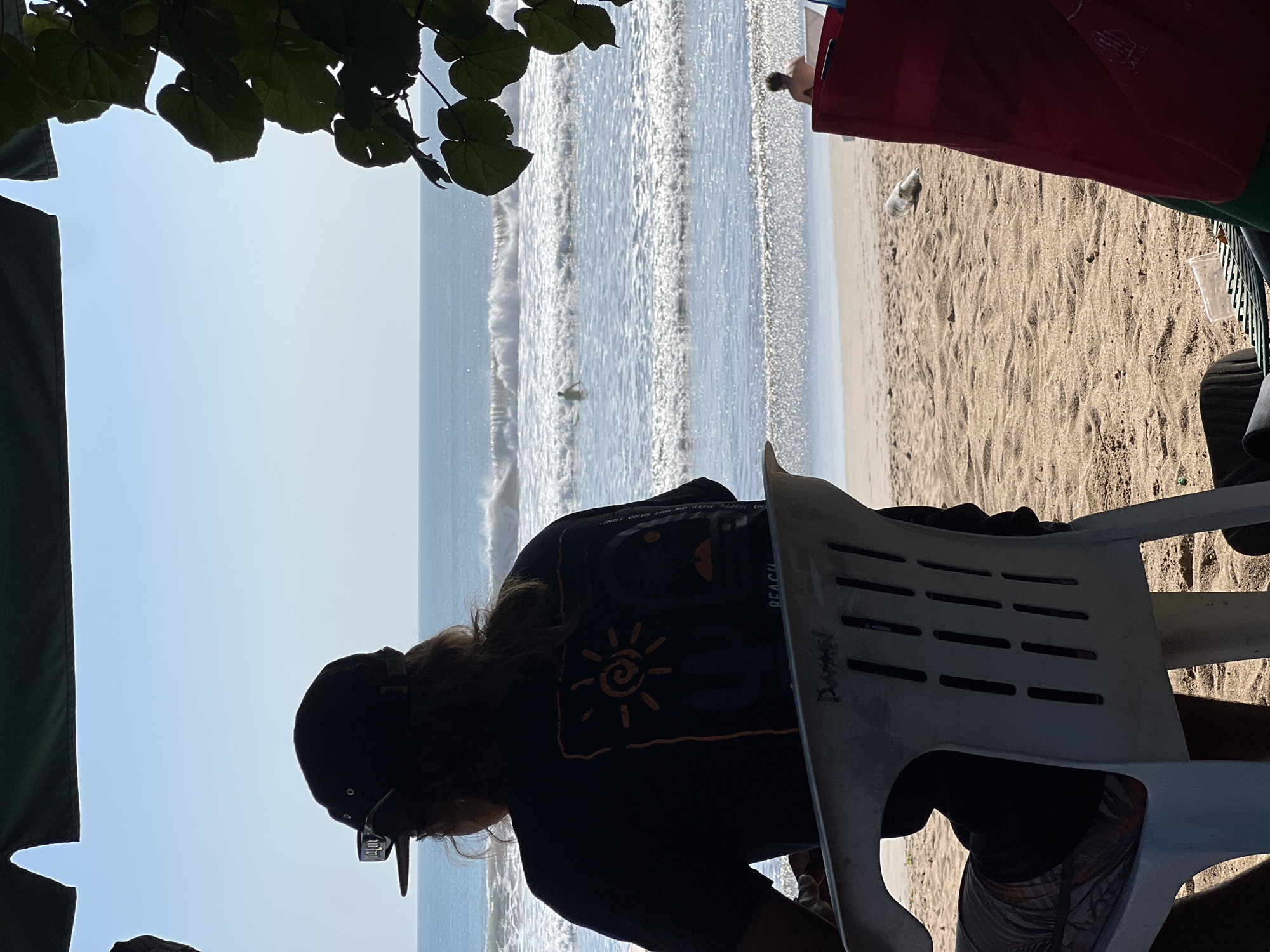 A person wearing a black baseball cap and a black t-shirt with an orange sun graphic on the back sits in a white plastic chair facing a beach and the ocean. The person's legs are crossed and they are looking out at the water. Waves are crashing on the shore, and a surfer is visible in the distance. The sand is light brown, and there are some green leaves in the foreground on the right side of the image. The sky is clear and blue. The scene appears to be in Kuta, Indonesia, based on the common beach activities and scenery depicted.