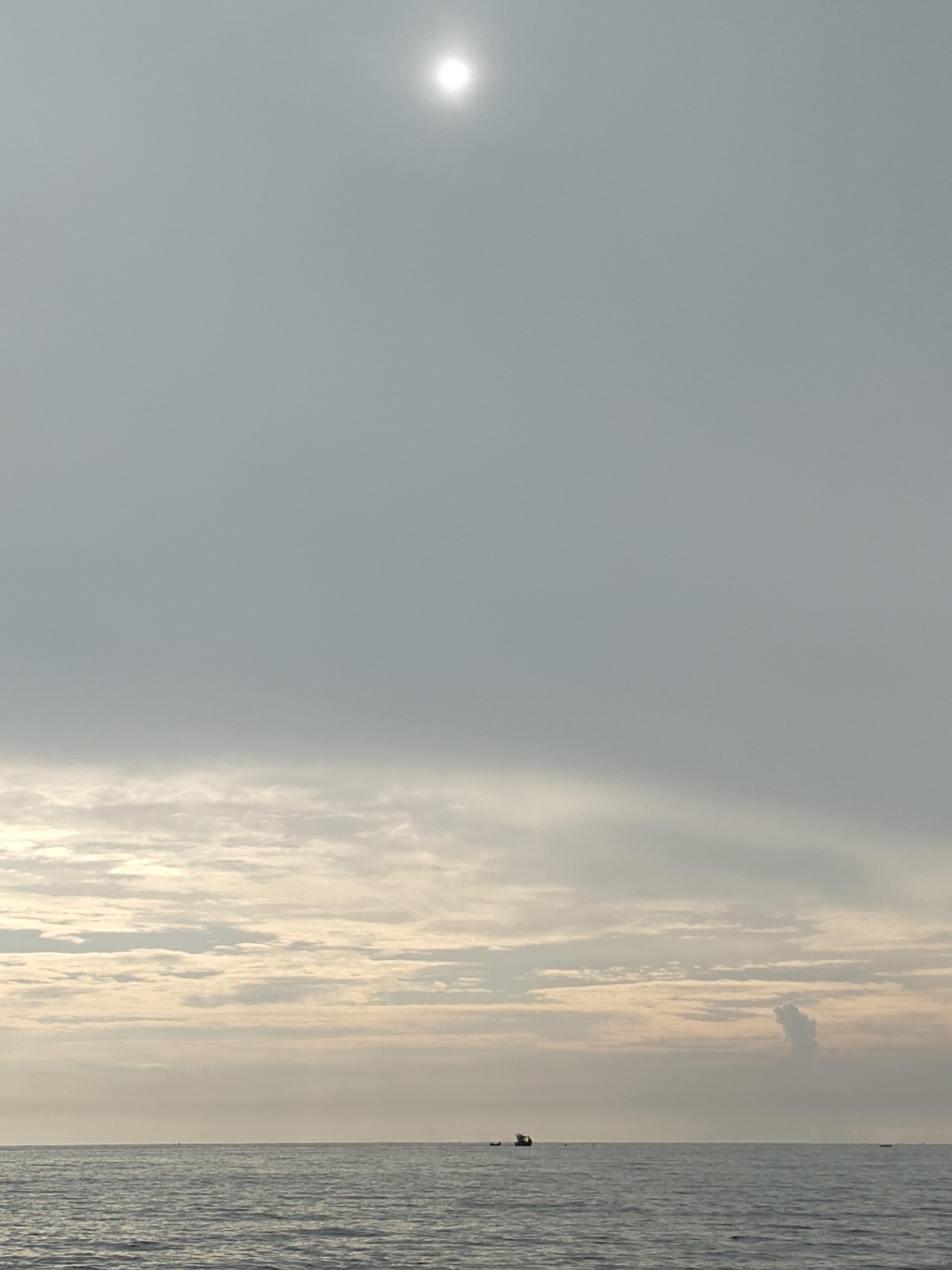 The image depicts a serene seascape at what appears to be either sunrise or sunset. The sky is a pale grey with subtle hints of pink and yellow near the horizon, suggesting soft lighting conditions. A bright, diffused sun is visible in the upper portion of the frame, its light scattering through the atmospheric haze. Wispy clouds are scattered across the sky, forming a textured layer just above the sea.
The sea itself is a deep blue-grey, with gentle ripples indicating calm waters. In the distance, several small boats are visible on the horizon. One boat, to the left, is a dark silhouette with a discernible structure, possibly a fishing vessel. Another even smaller boat is visible further to the right. There is a peculiar cloud formation to the right of the frame, resembling a vertical, somewhat amorphous shape.
The image is likely taken from the coast of Lhokseumawe, Indonesia, a city known for its coastline and proximity to the sea. The overall impression is one of tranquility and vastness, with the human presence indicated only by the distant boats. There are no people directly visible in the frame. The environment is natural, dominated by the sky and the sea.