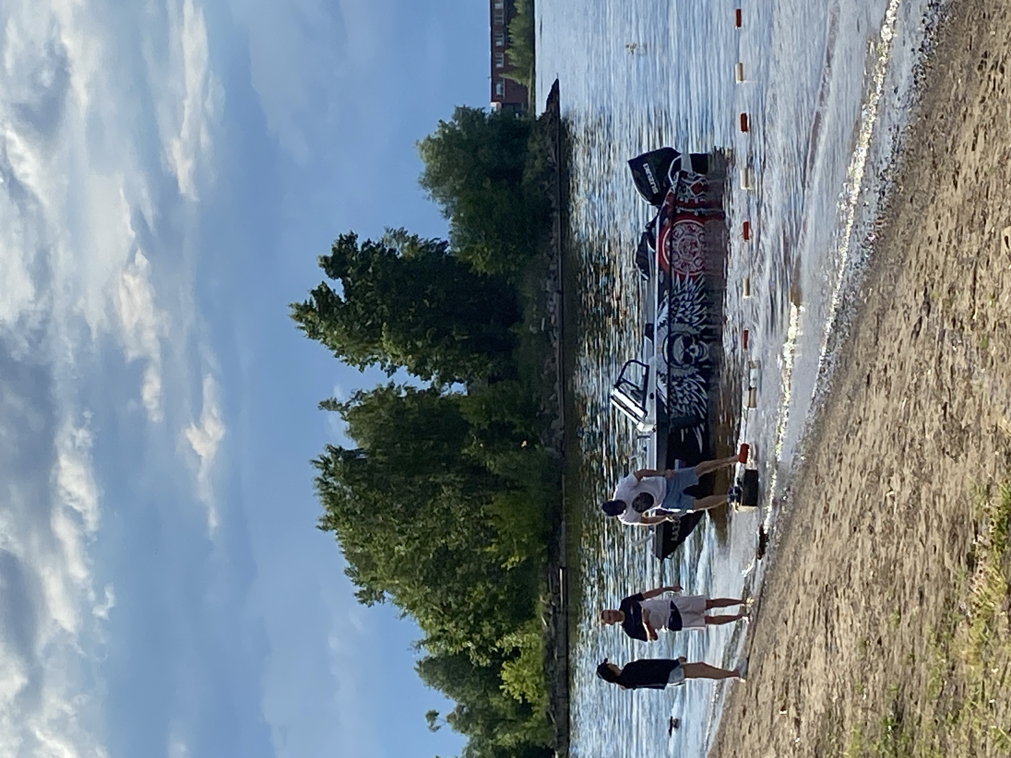 Three people, two women and one man, stand on a sandy shore next to a body of water. A man is in the water next to a boat, appearing to be preparing it for launch or maneuvering it. The boat has a graphic design on its side featuring skulls and wings. In the background, a small island with dense green trees is visible, and beyond that, buildings are seen on the horizon. The sky is cloudy and overcast. The scene appears to be a recreational area near Chaikovsky, Russia, possibly a boat launch or a beach area along the Kama River reservoir.