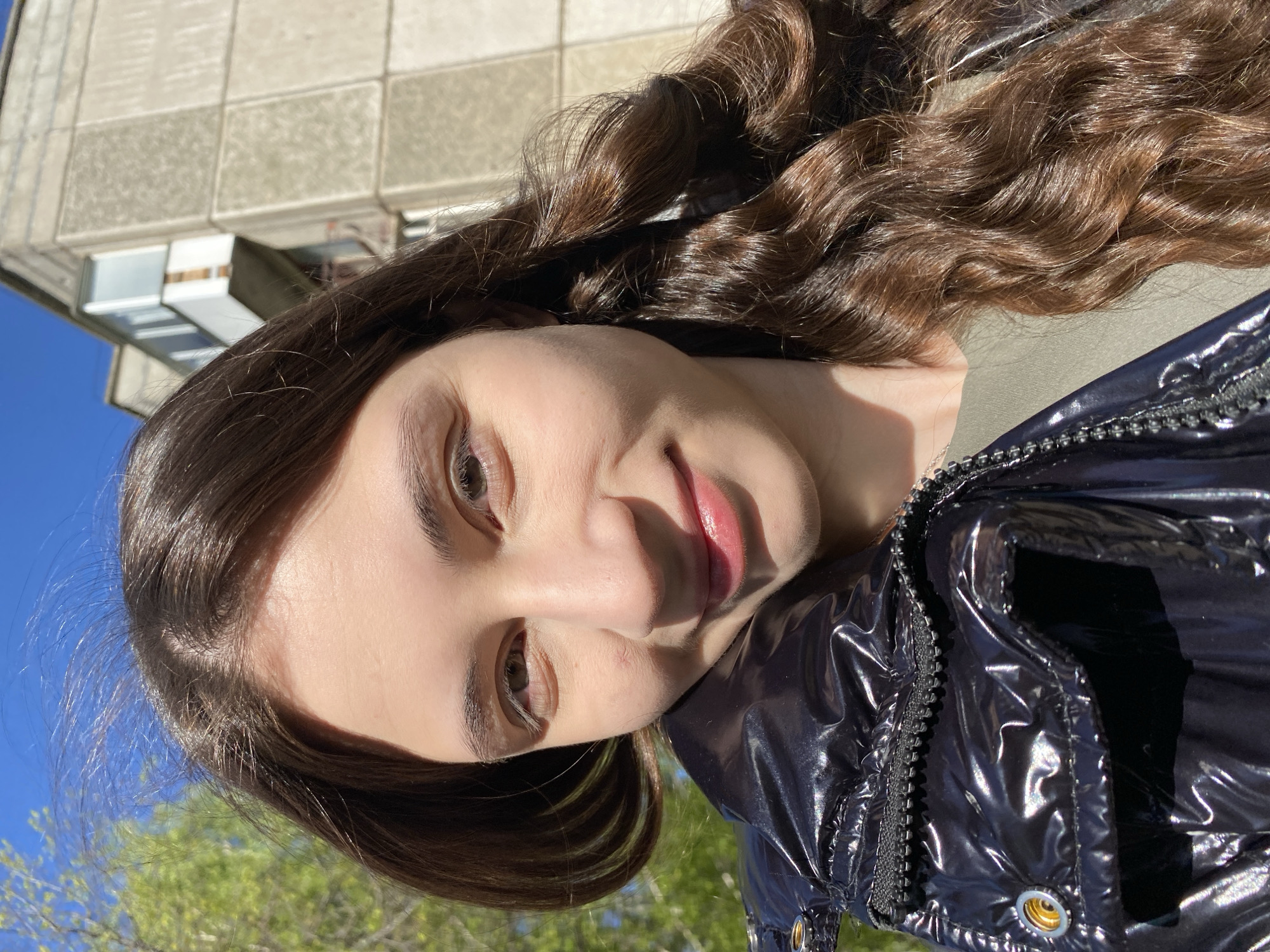 A close-up, slightly angled selfie of a woman with long, wavy brown hair. She has light skin and is wearing a shiny, dark jacket. The background shows a building made of concrete blocks, possibly an apartment building, along with a glimpse of blue sky and green trees. It is daytime and the lighting is bright, suggesting a sunny day. There is no explicit indication that the location is Votkinsk, Russia.