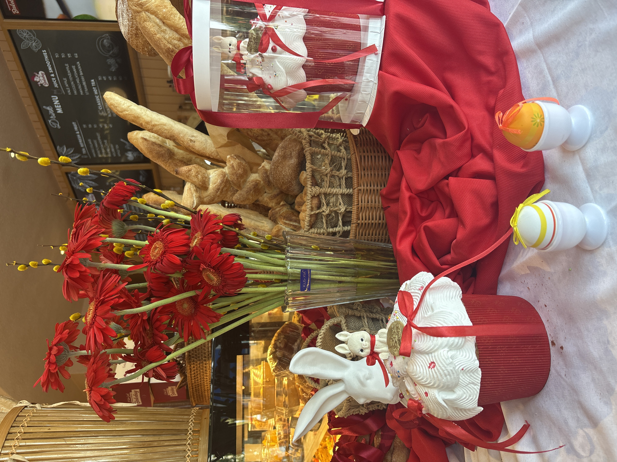 The image shows a display of Easter decorations and baked goods in what appears to be a bakery or cafe. There are loaves of bread and other pastries arranged in baskets. A tall vase of red gerbera daisies and yellow pussy willow branches is positioned at the front. Several Easter-themed decorations are present, including rabbit figurines, decorative eggs, and a wrapped panettone-like cake with white icing and a red ribbon. The display is set against a backdrop of red fabric and a white tablecloth. Behind the display, there's a menu board with "Drink Menu" and listings of juices, smoothies, and teas. A portion of the bakery's interior is visible in the reflection of the glass case. The location appears to be in Bắc Nha Trang, Vietnam, based on the bakery name "Bakebry" visible in the top left of the picture and "DELISOGA" shown in the vase of red gerberas.
