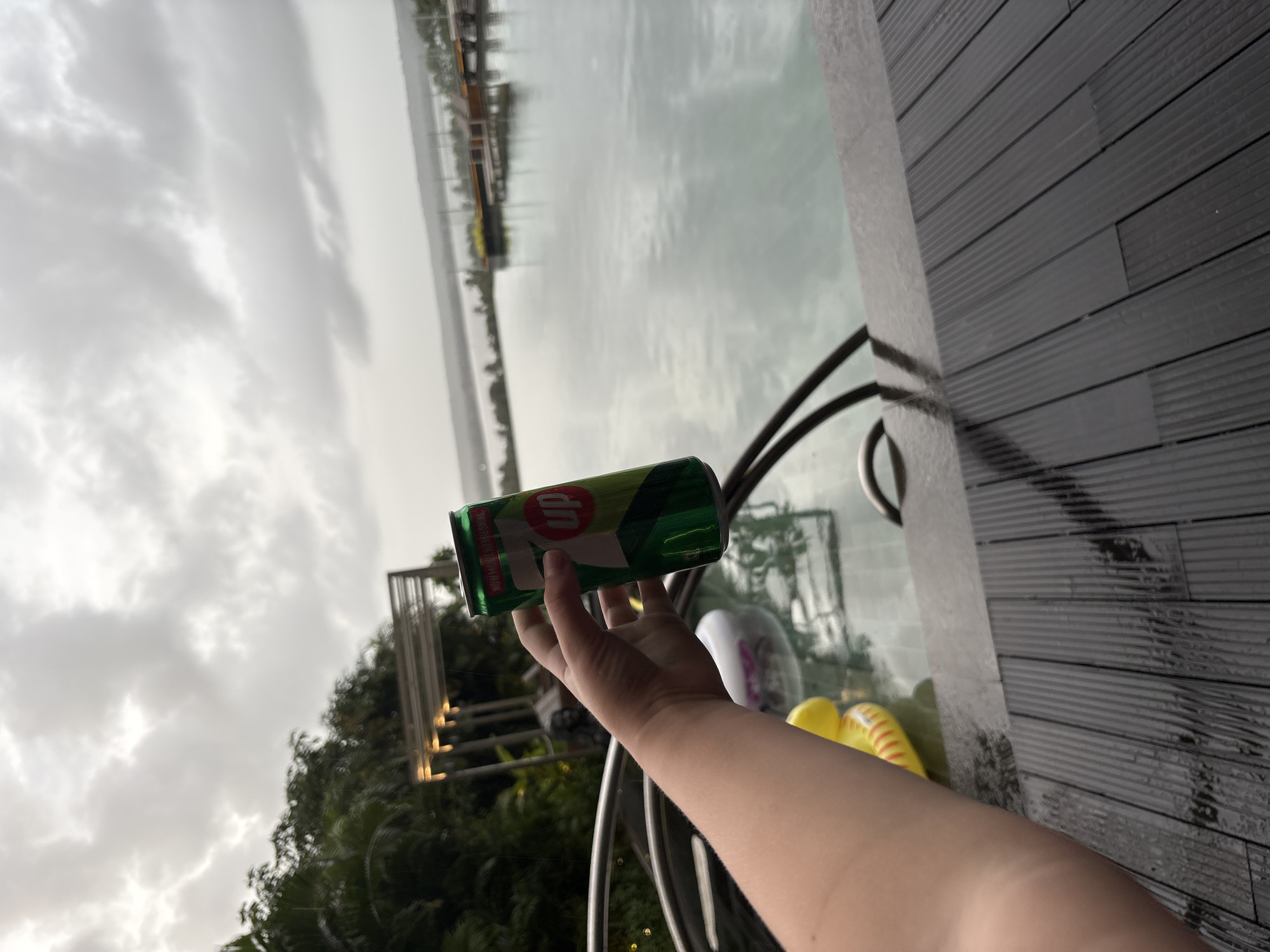 A person's arm and hand are holding a green 7UP can above a wet, dark wooden deck and a swimming pool. The pool extends to the horizon, where a calm body of water is visible under a cloudy sky. In the background, there are trees and structures with lights. The overall impression is one of a resort or hotel setting. The image was taken in Vagator, India.