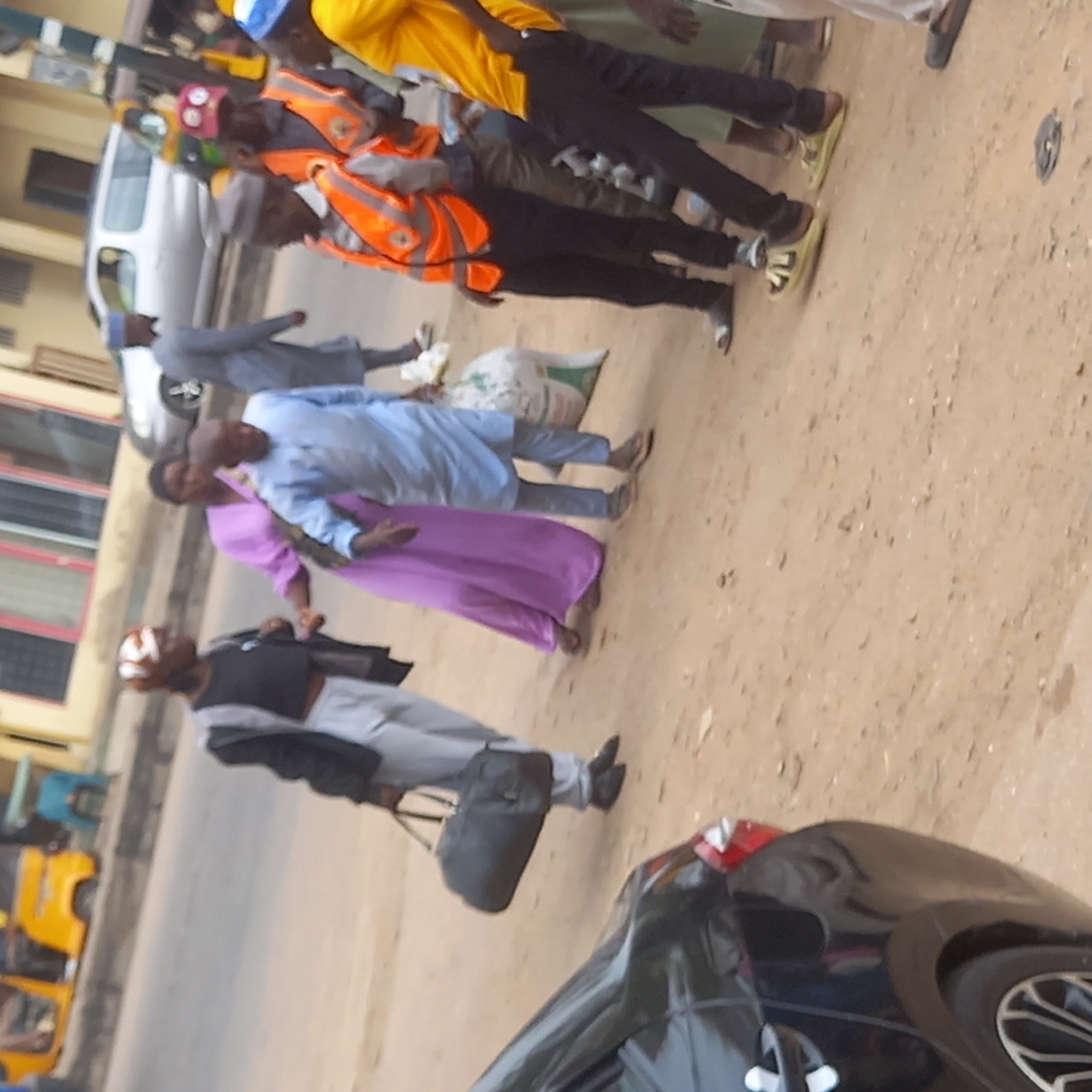 A street scene in Dadi Riba, Nigeria shows a group of people walking on a dirt-covered sidewalk alongside a road.
There are at least eight individuals visible. One man wears a yellow shirt and blue baseball cap. Another man is wearing an orange safety vest and a hat with red markings. A man in a light blue shirt and trousers walks beside a woman in a purple dress. Another man with bandages around his head, a black vest, and gray trousers carries a dark bag.
A white van, a yellow auto rickshaw, and a black car are visible near the street. A bag is on the sidewalk. Buildings are in the background.